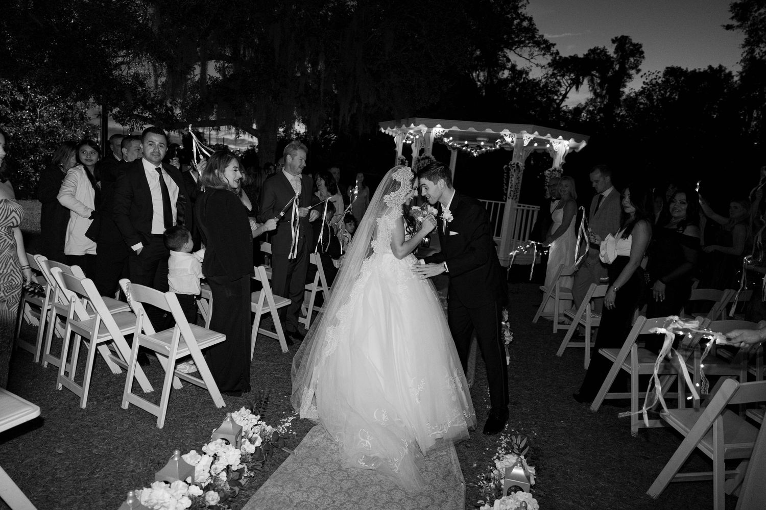 Outdoor evening wedding ceremony with white chairs and guests.
