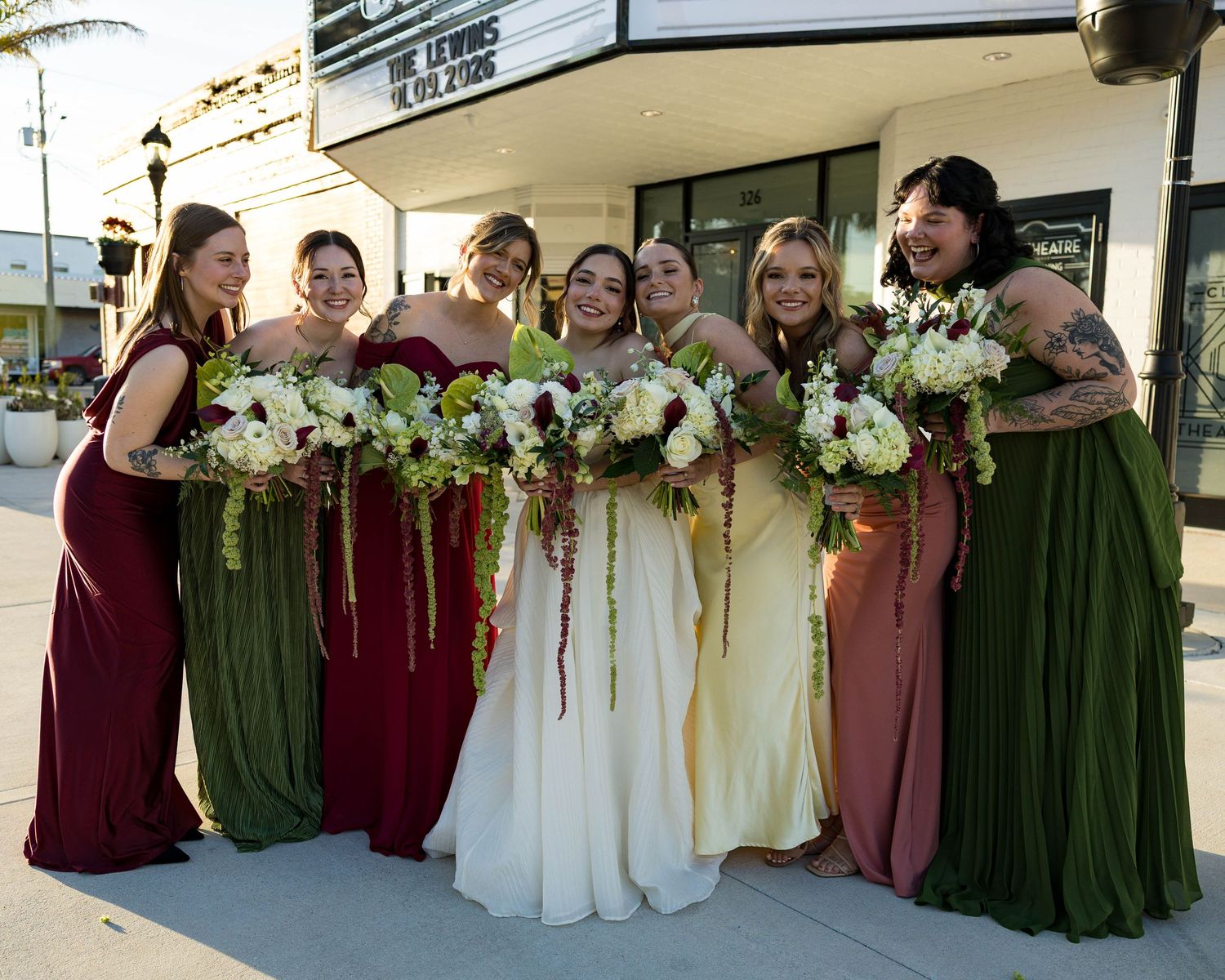 Wedding party poses outside a historic theater in burgundy, green and cream colored dresses.