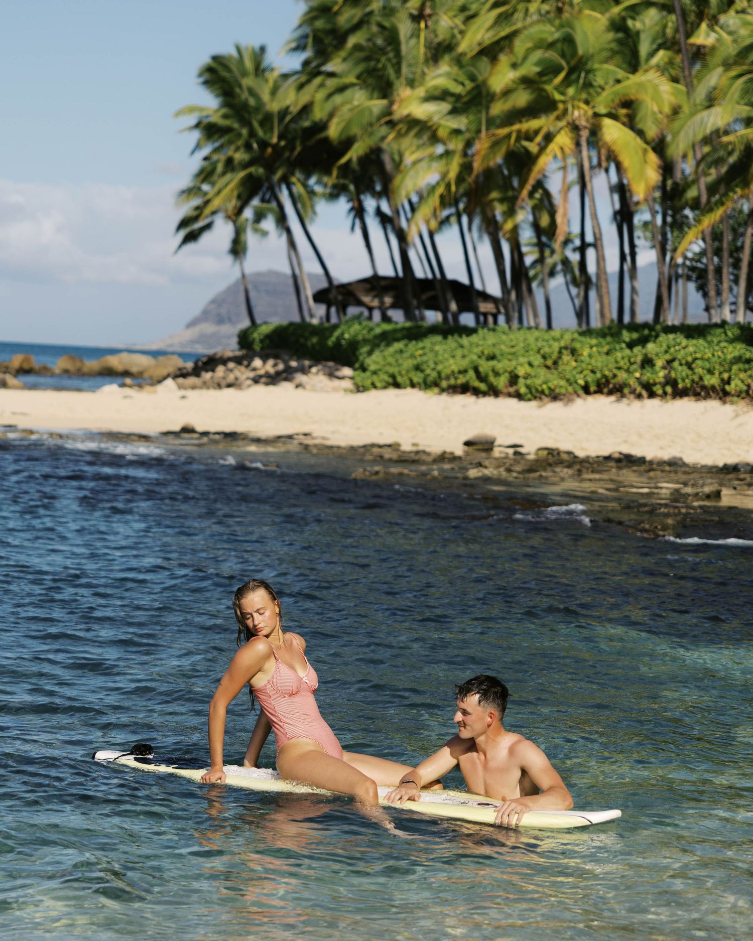 Two people relax on a surfboard in crystal clear tropical waters near a palm-lined beach in Hawaii.