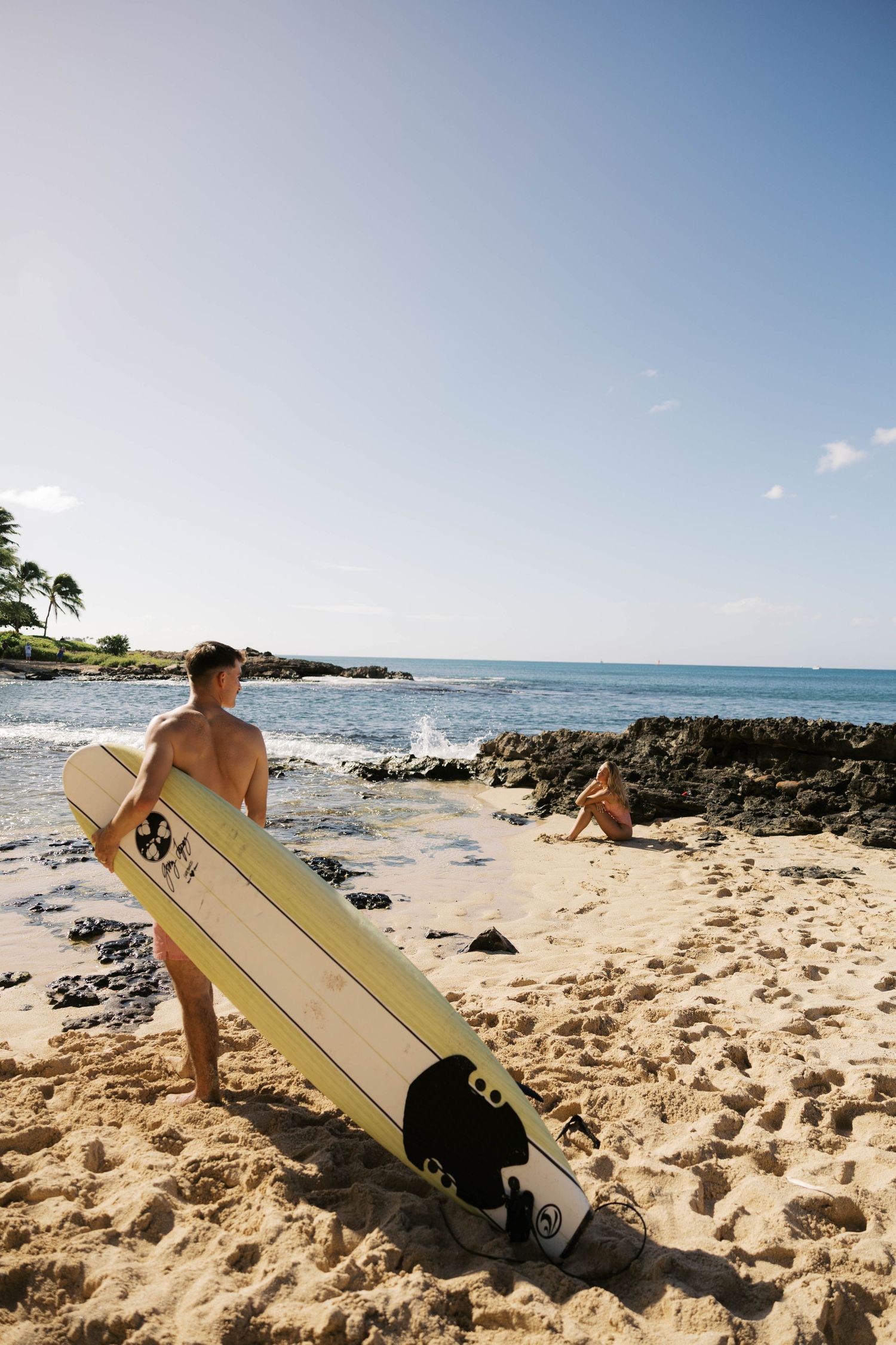 Surfer holding longboard stands on rocky beach with ocean waves and blue sky in Hawaii.