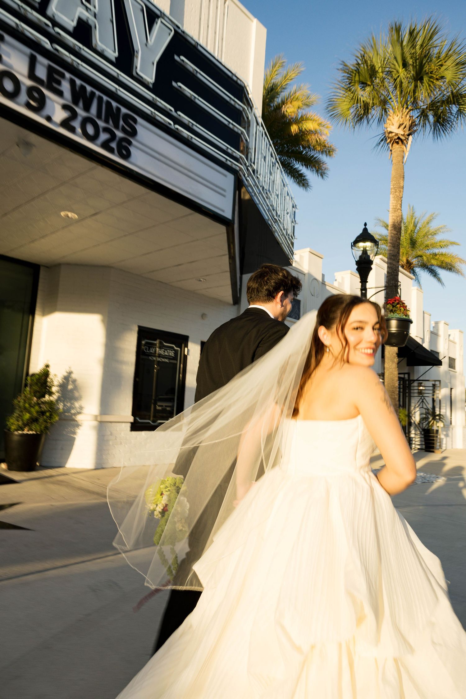 A bride in a flowing white gown and veil poses outside a theater with palm trees in the background.