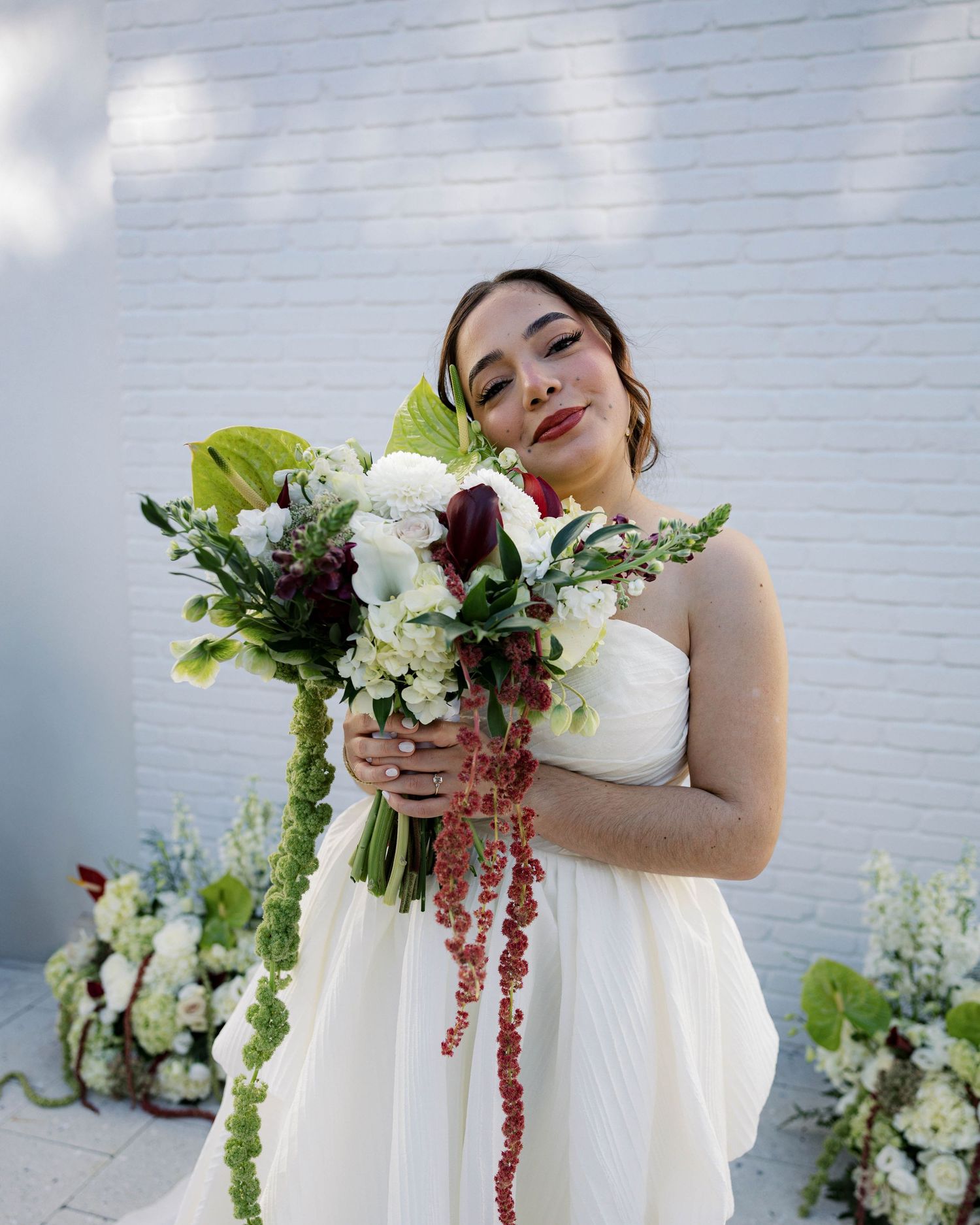 Bride holding a lush bouquet with cascading flowers against a white brick wall.