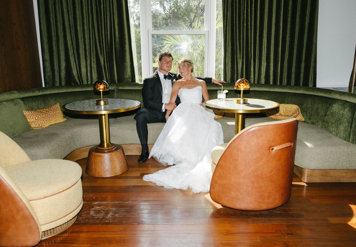 Bride and groom sitting together in an elegant green velvet lounge with gold accent tables on their wedding day.