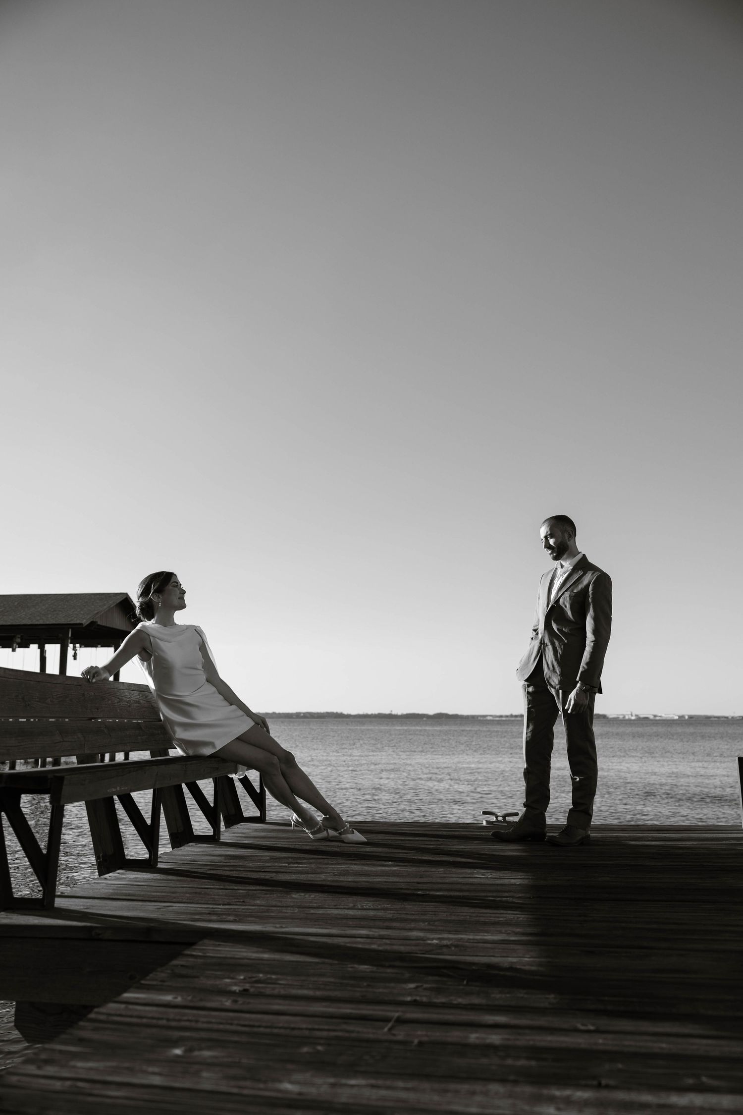 Silhouettes of two people on a wooden pier against a bright sky.