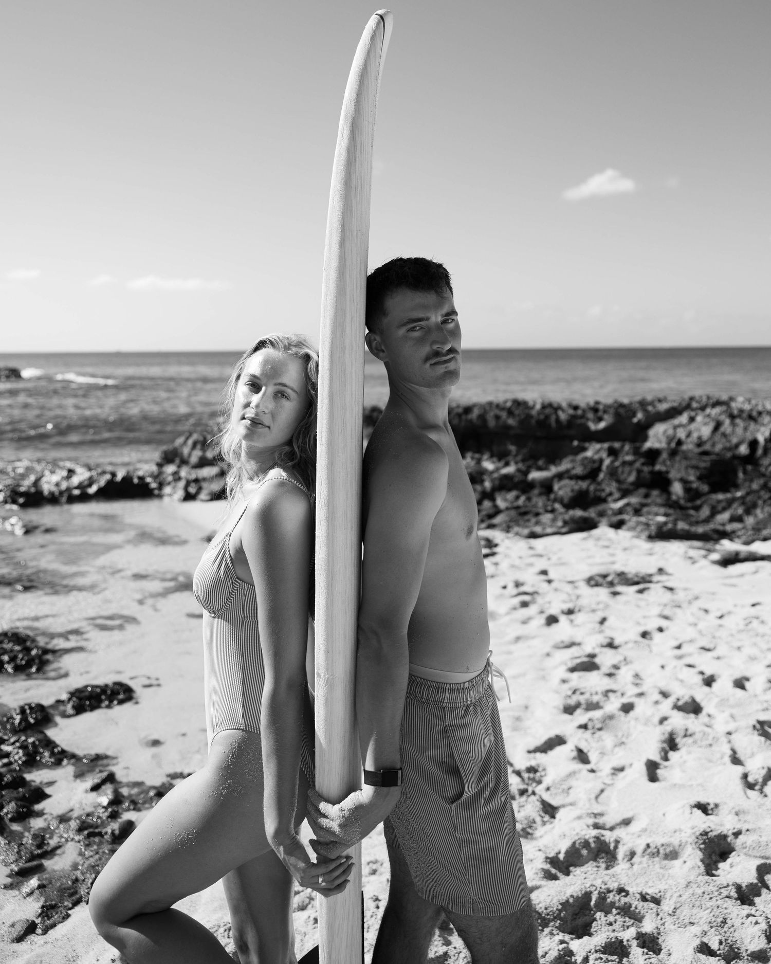 Black and white beach portraits with surfboard against rocky shoreline.