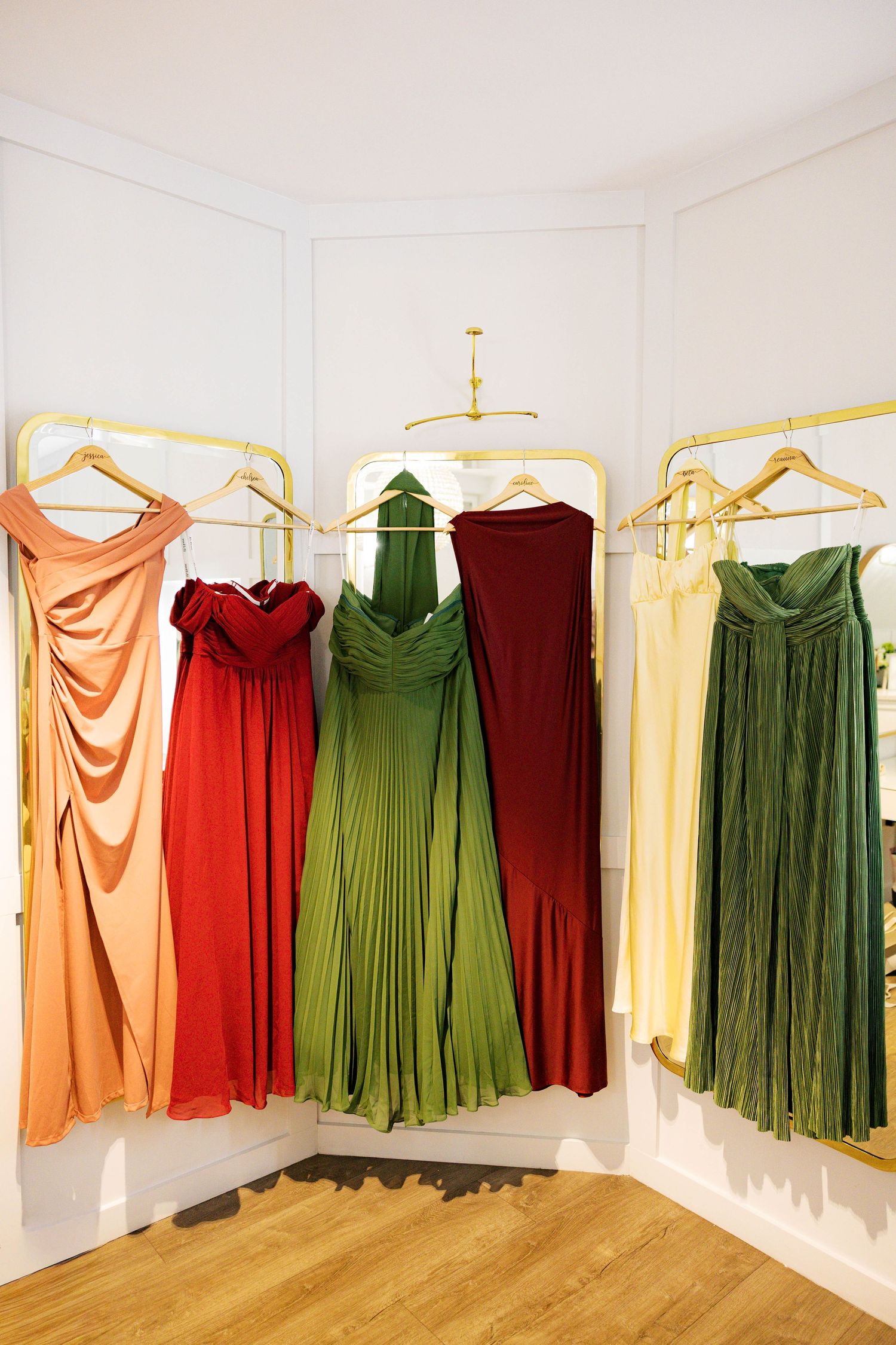 Collection of formal dresses in green, red, and peach tones displayed on gold hangers against white walls.