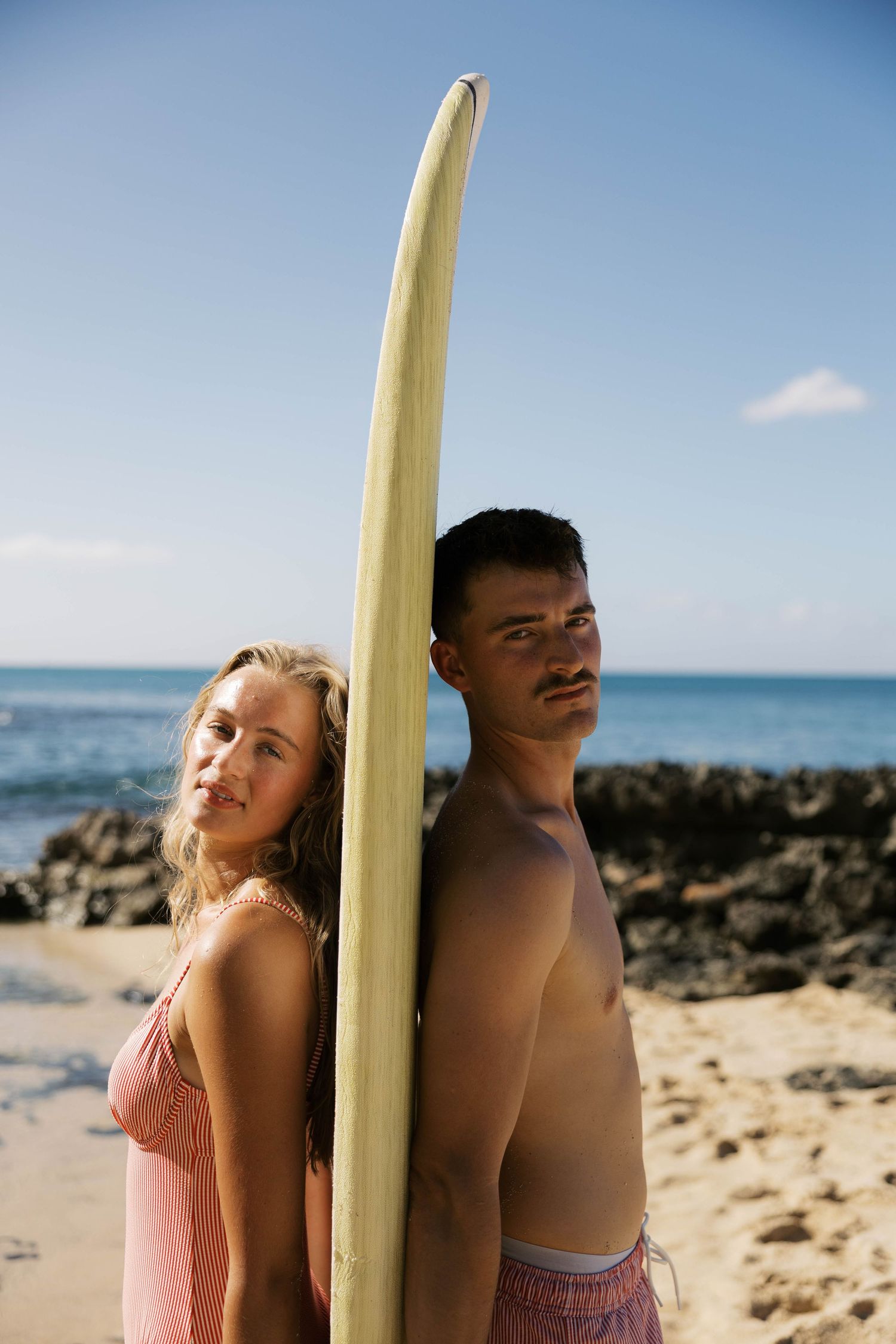 Couple posing with a surfboard on a sunny beach with ocean waves in the background.