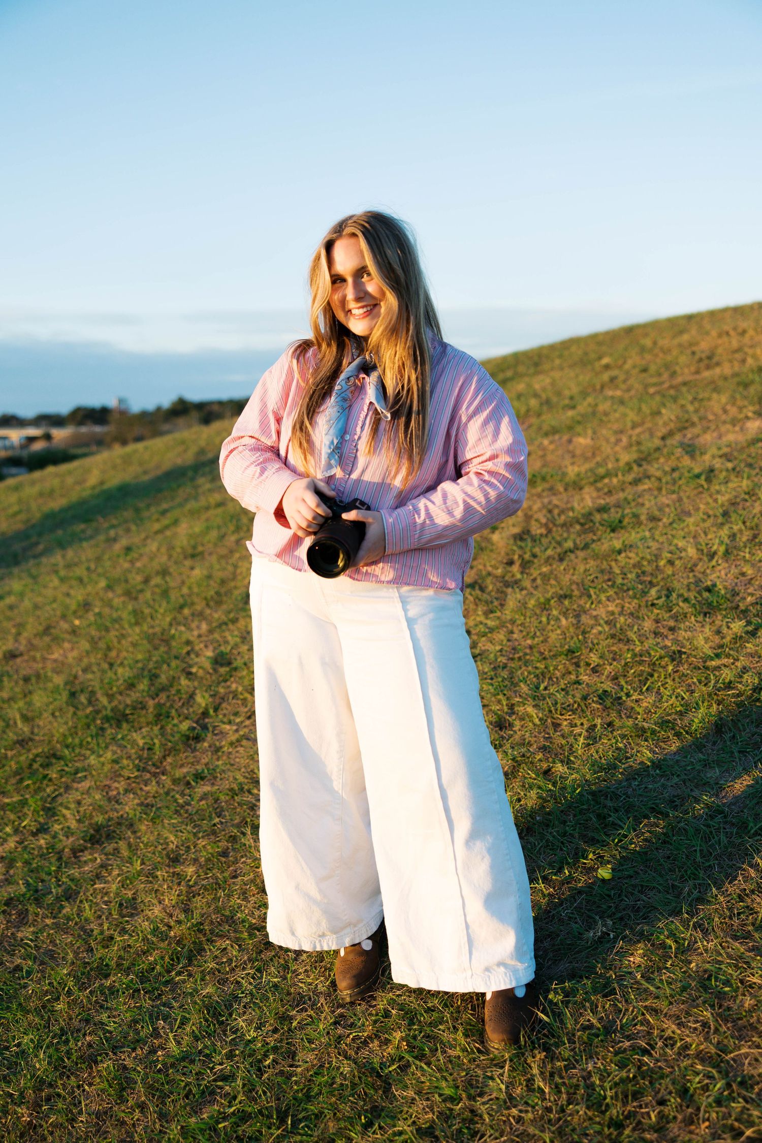 Someone wearing a pink jacket and white dress sits on a grassy hillside during golden hour.
