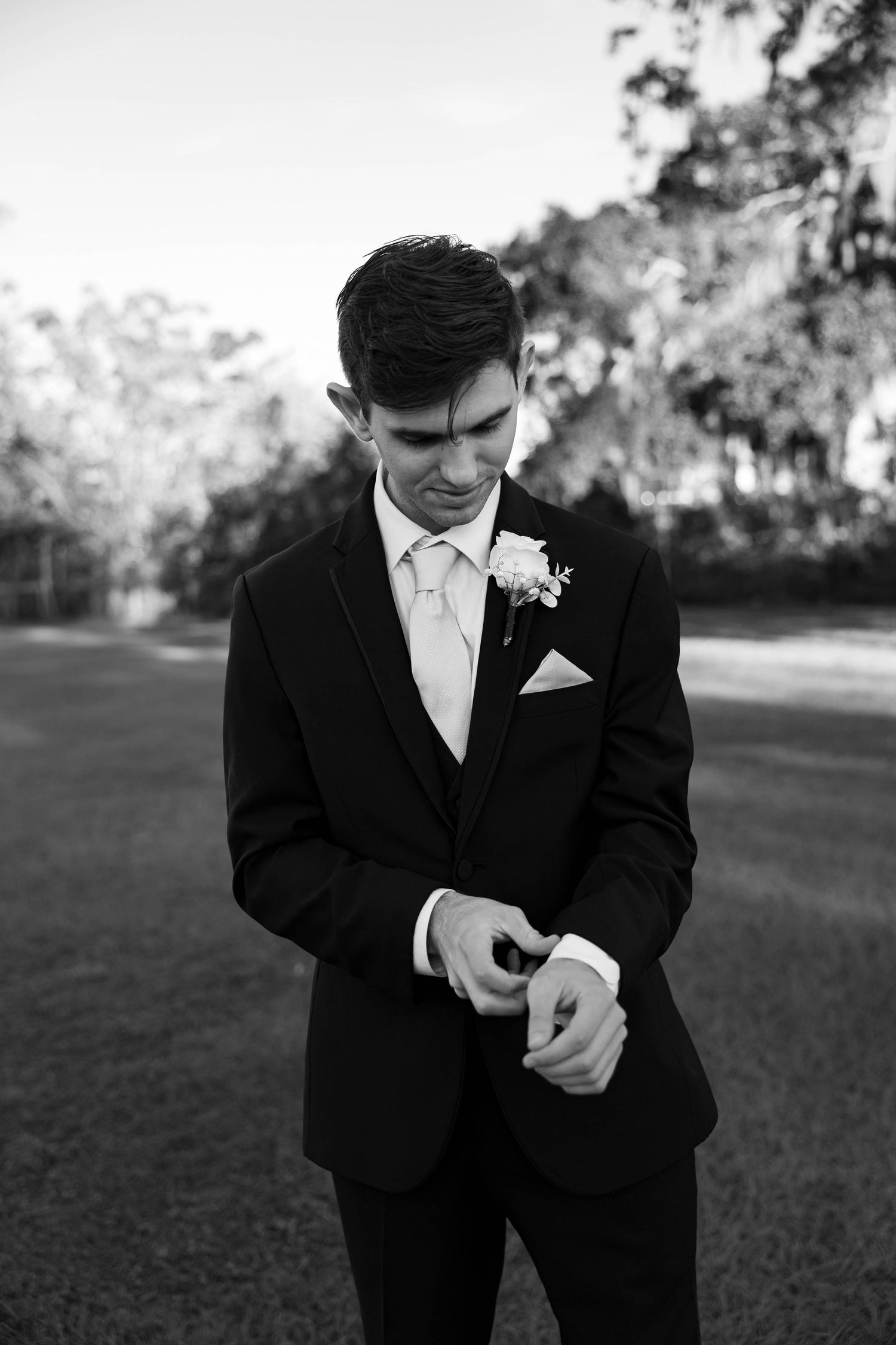 Black and white photo of groom adjusting his cufflinks outdoors.