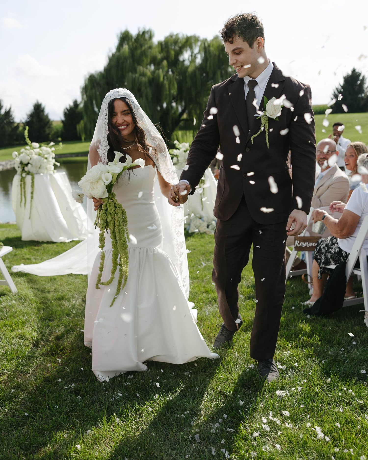 Joyful newlywed couple walking down outdoor aisle as guests toss white flower petals at wedding ceremony.