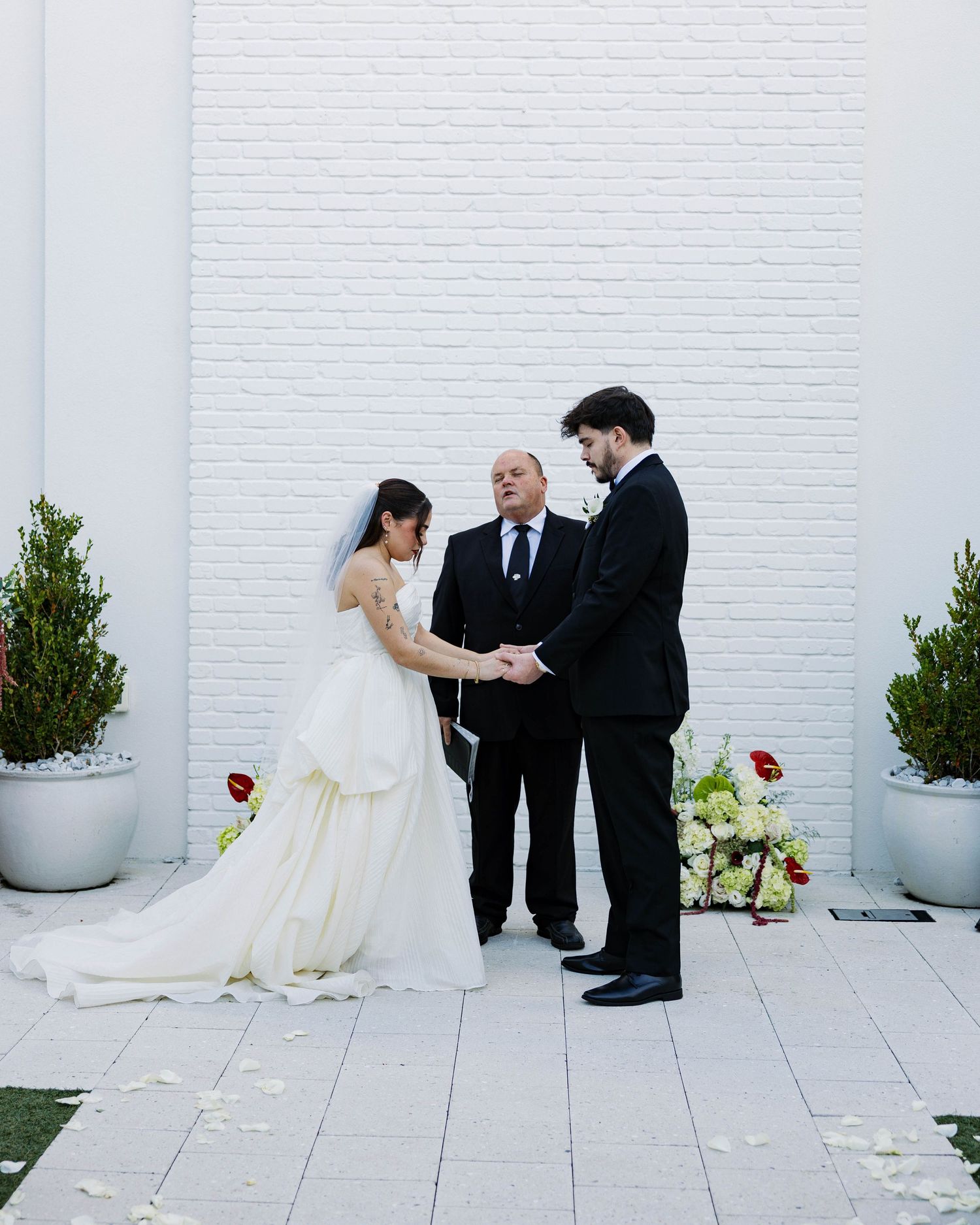 Intimate wedding ceremony moment against white brick wall with potted plants.
