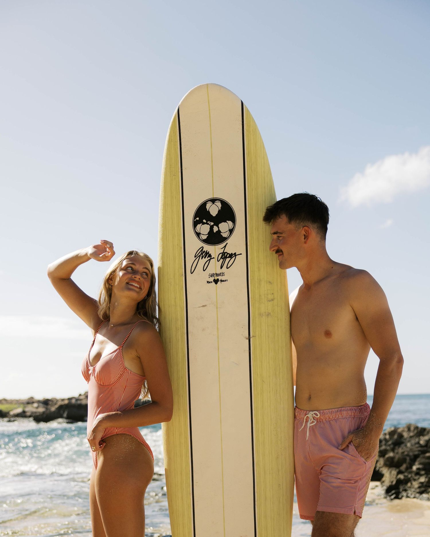 Joyful beach scene with two people posing beside cream surfboard on sunny day.