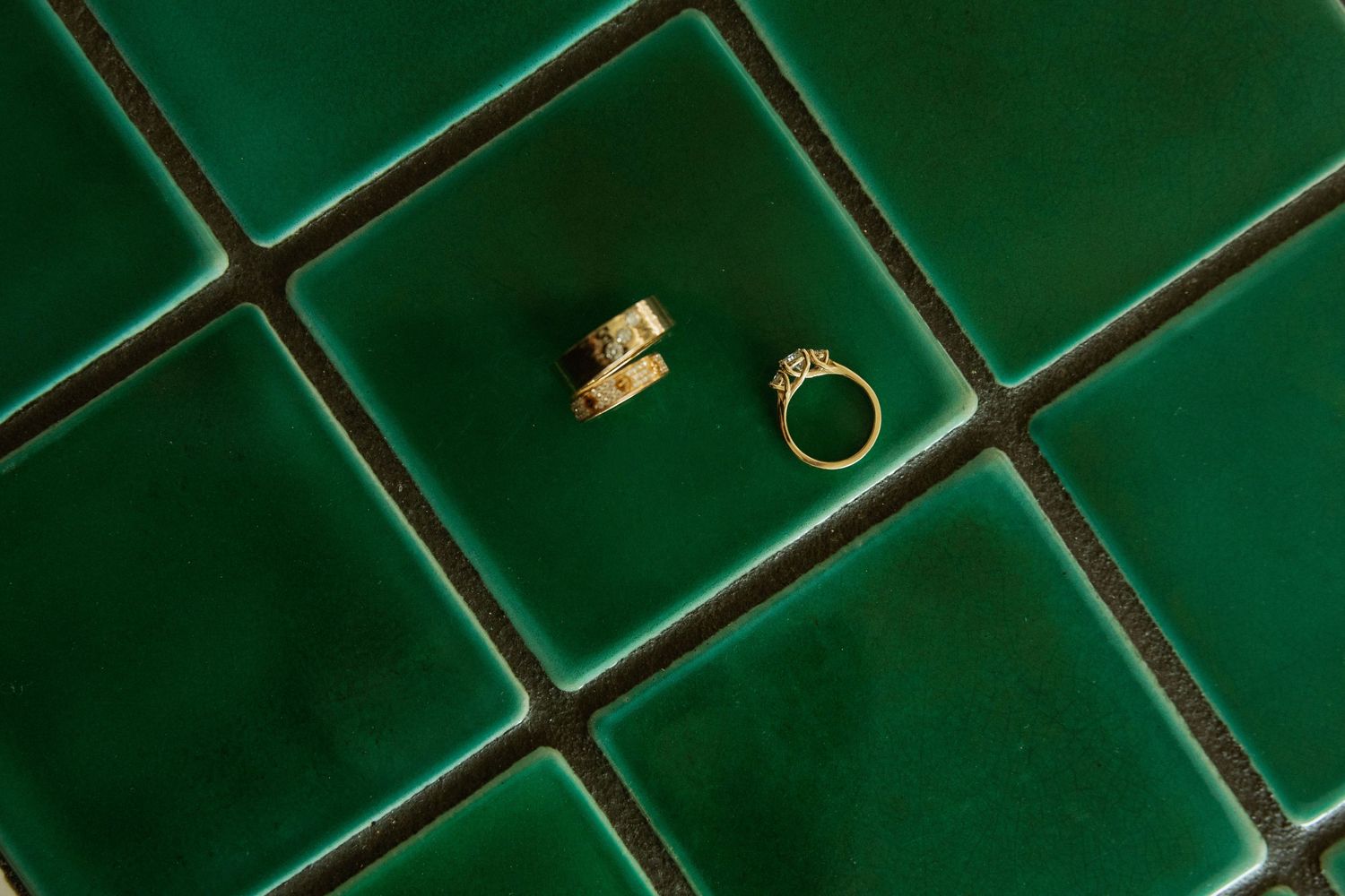 Two gold wedding rings, one band and one diamond engagement ring, displayed on green ceramic tiles.