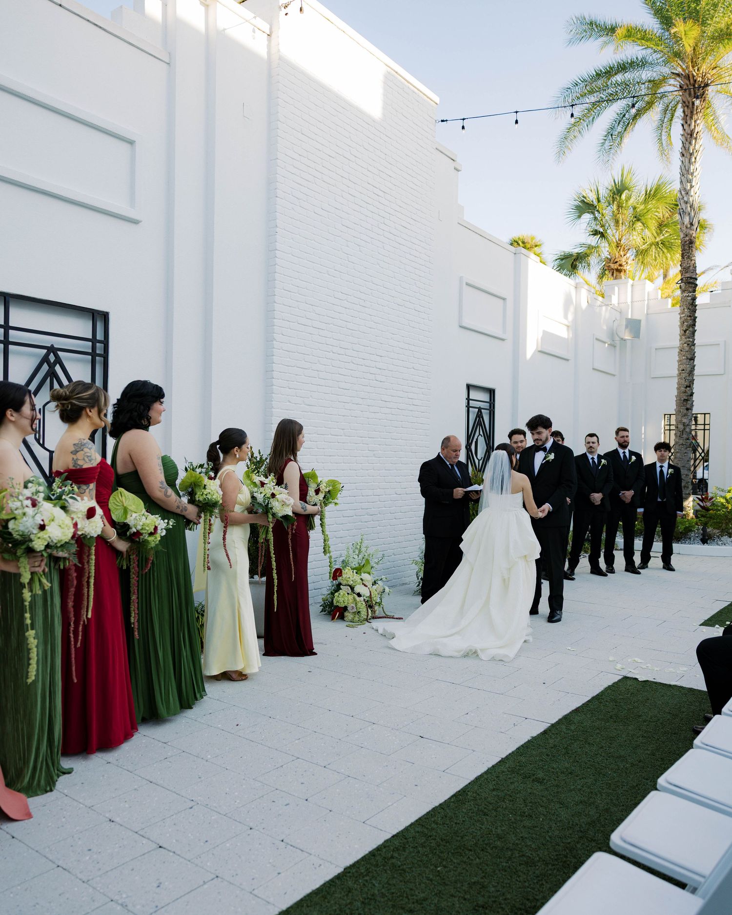Wedding ceremony setup with palm trees and white walls featuring bridesmaids in burgundy and green dresses.