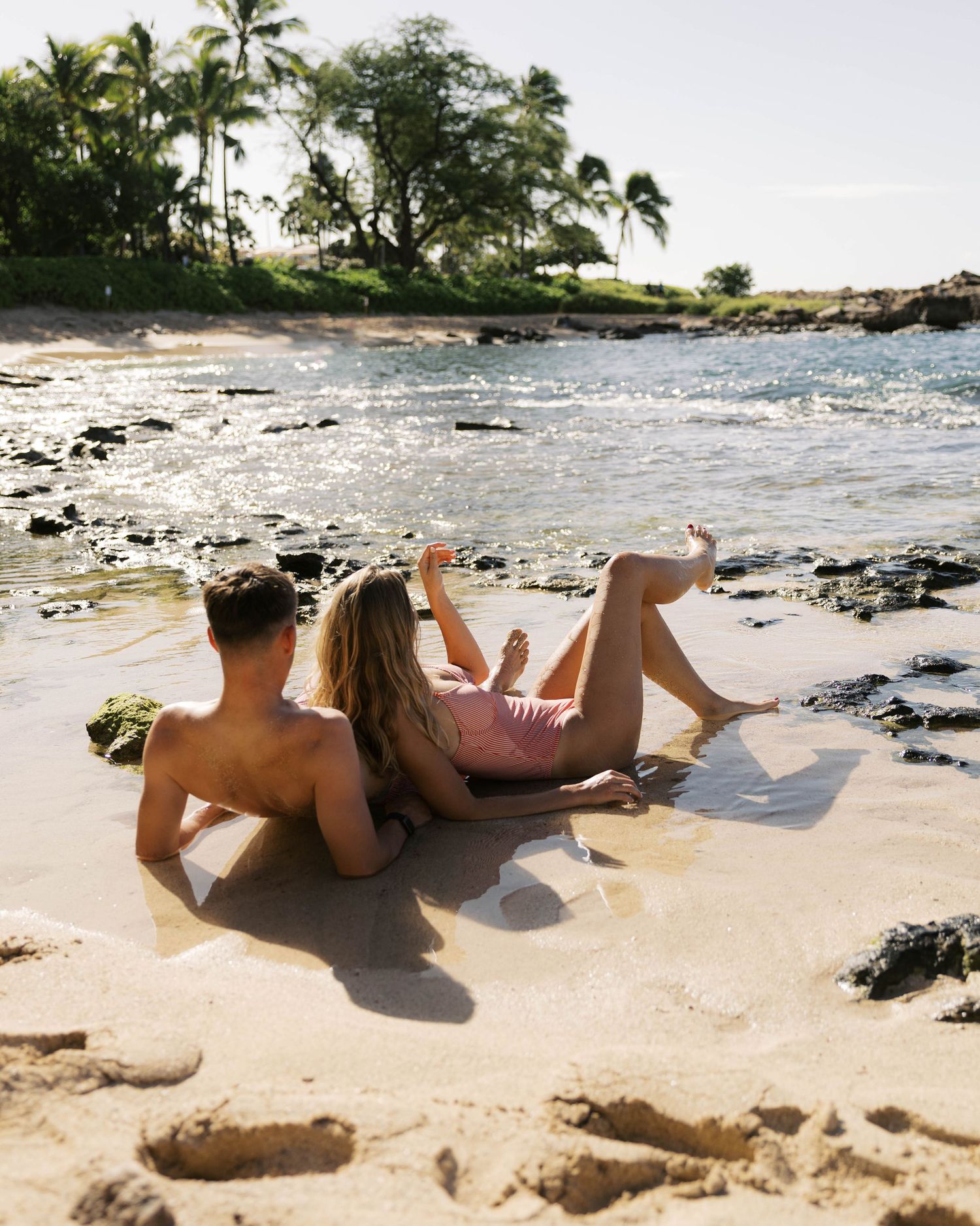 Wide angle view of beachgoer lounging on sandy tropical shore with volcanic rocks and palm trees.