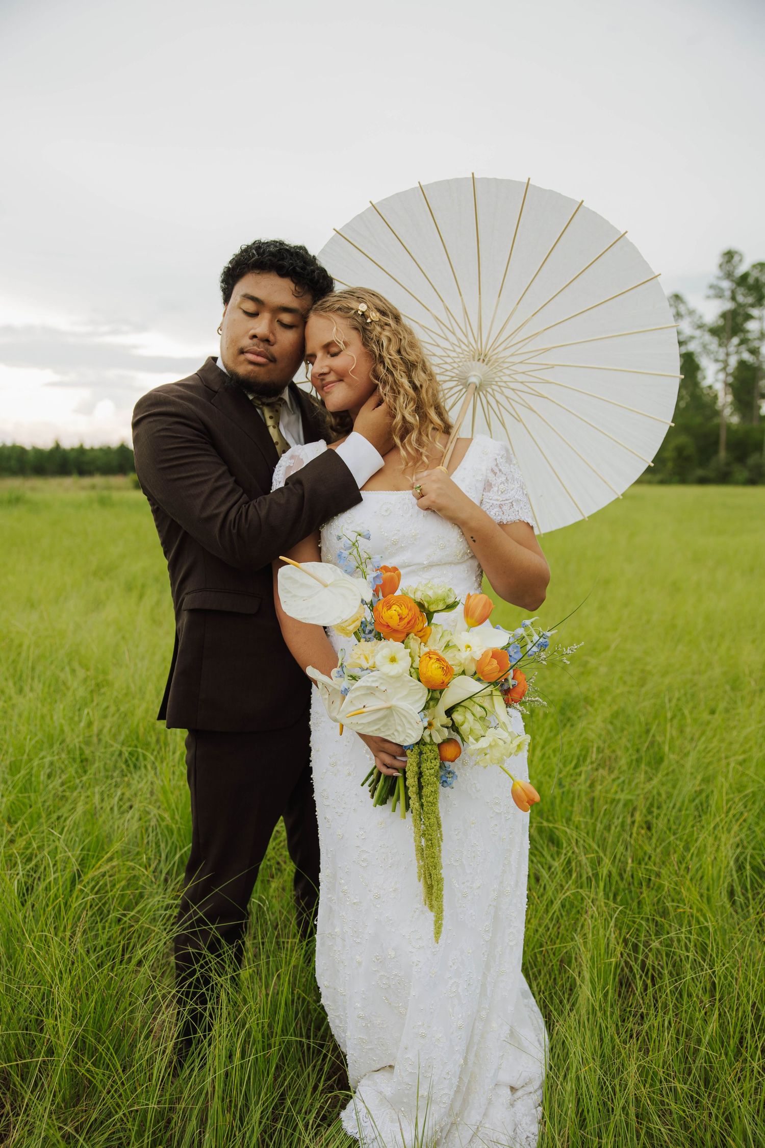 A couple shares romantic moments in a grassy field while holding a white parasol on their wedding day.