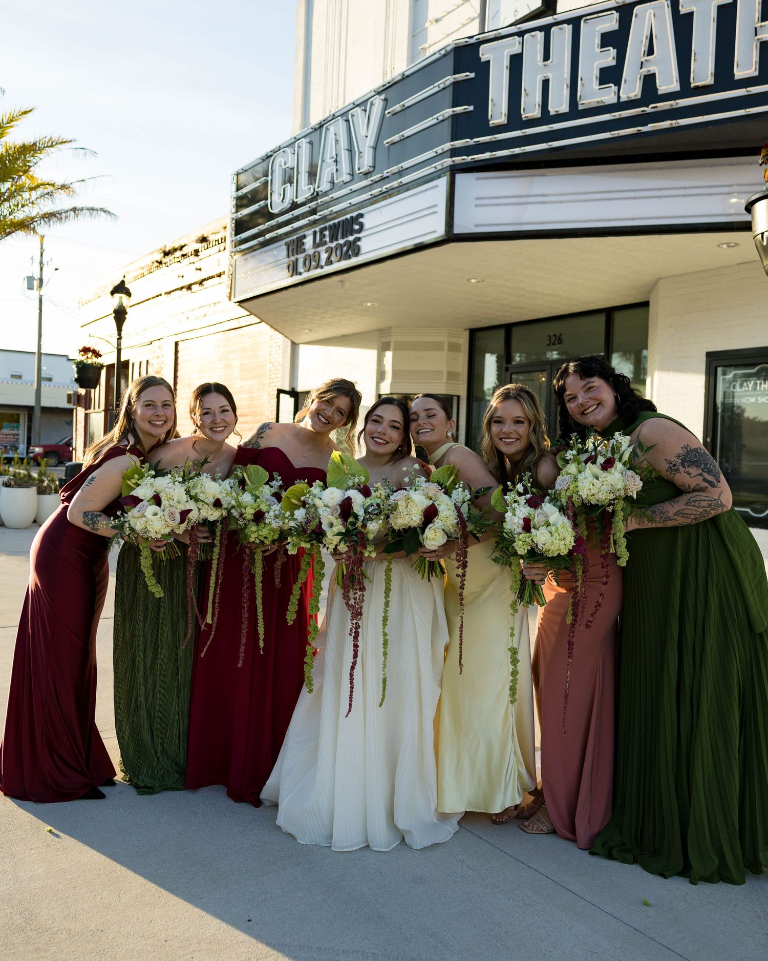 Wedding party arranged in formal line outside theater with bridesmaids in burgundy and green dresses.