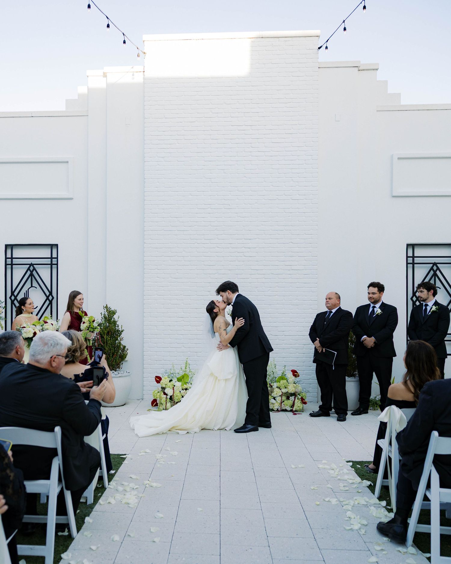 Outdoor wedding ceremony in progress with guests seated on white chairs against modern white architecture.