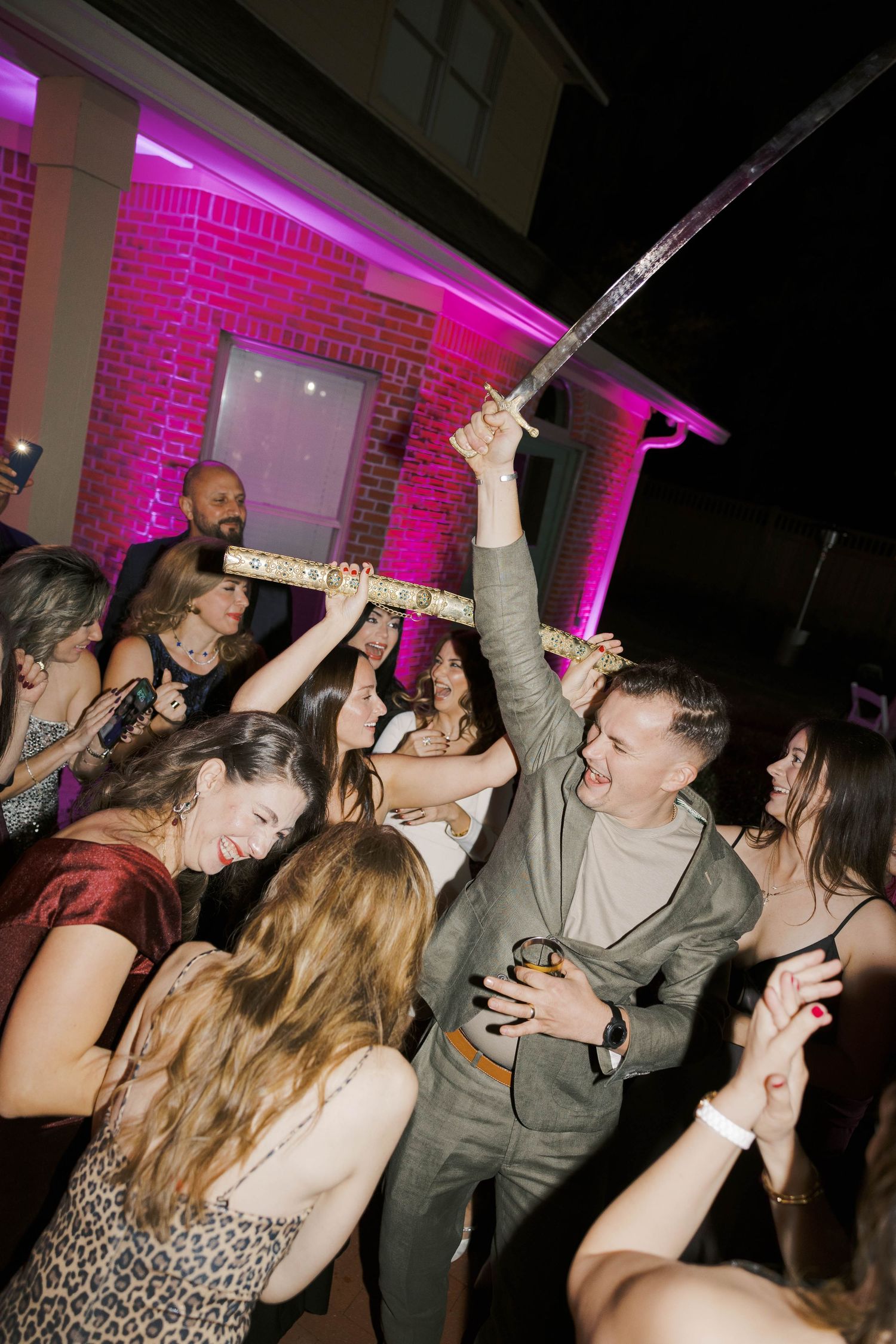 Wedding guests dancing and celebrating on a dance floor illuminated with pink lighting.