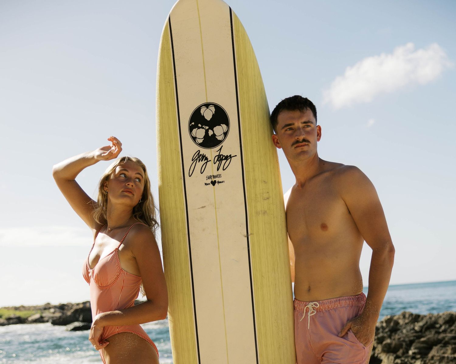 Playful beach photo with a couple and vintage-style surfboard against ocean backdrop.