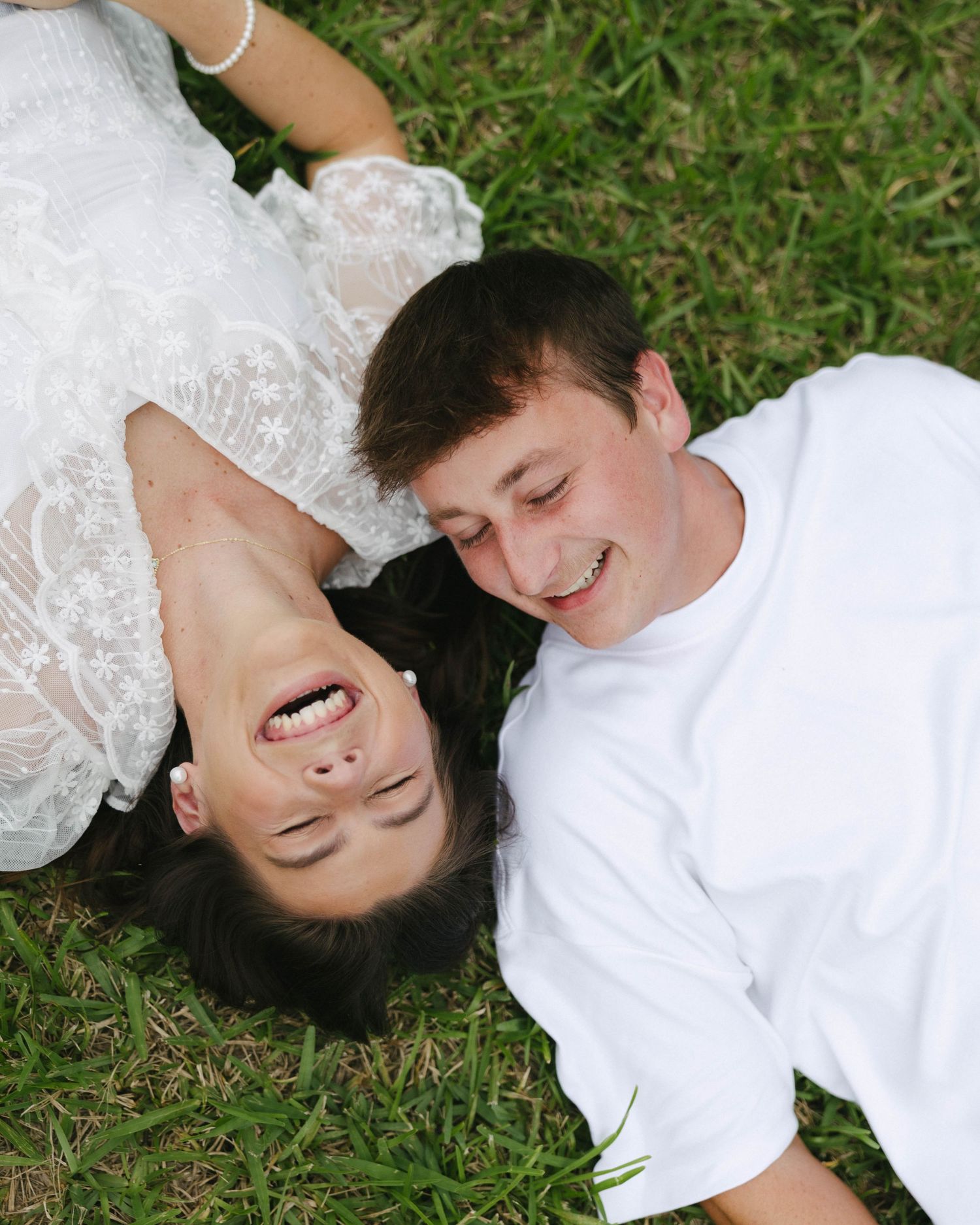 A joyful couple lying on green grass, smiling and laughing together, dressed in white outfits.