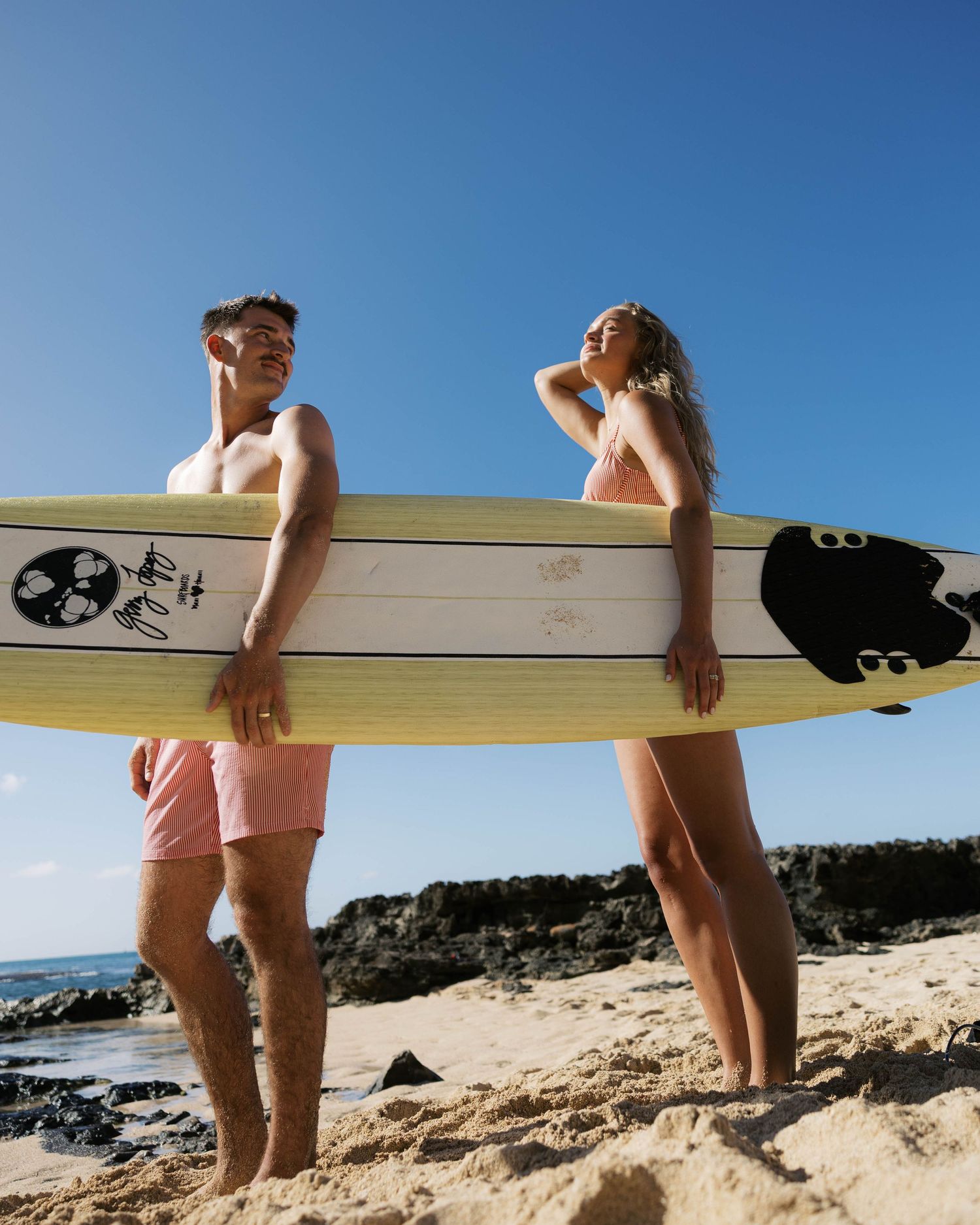 Two people holding a surfboard while walking on a sandy beach with ocean views.