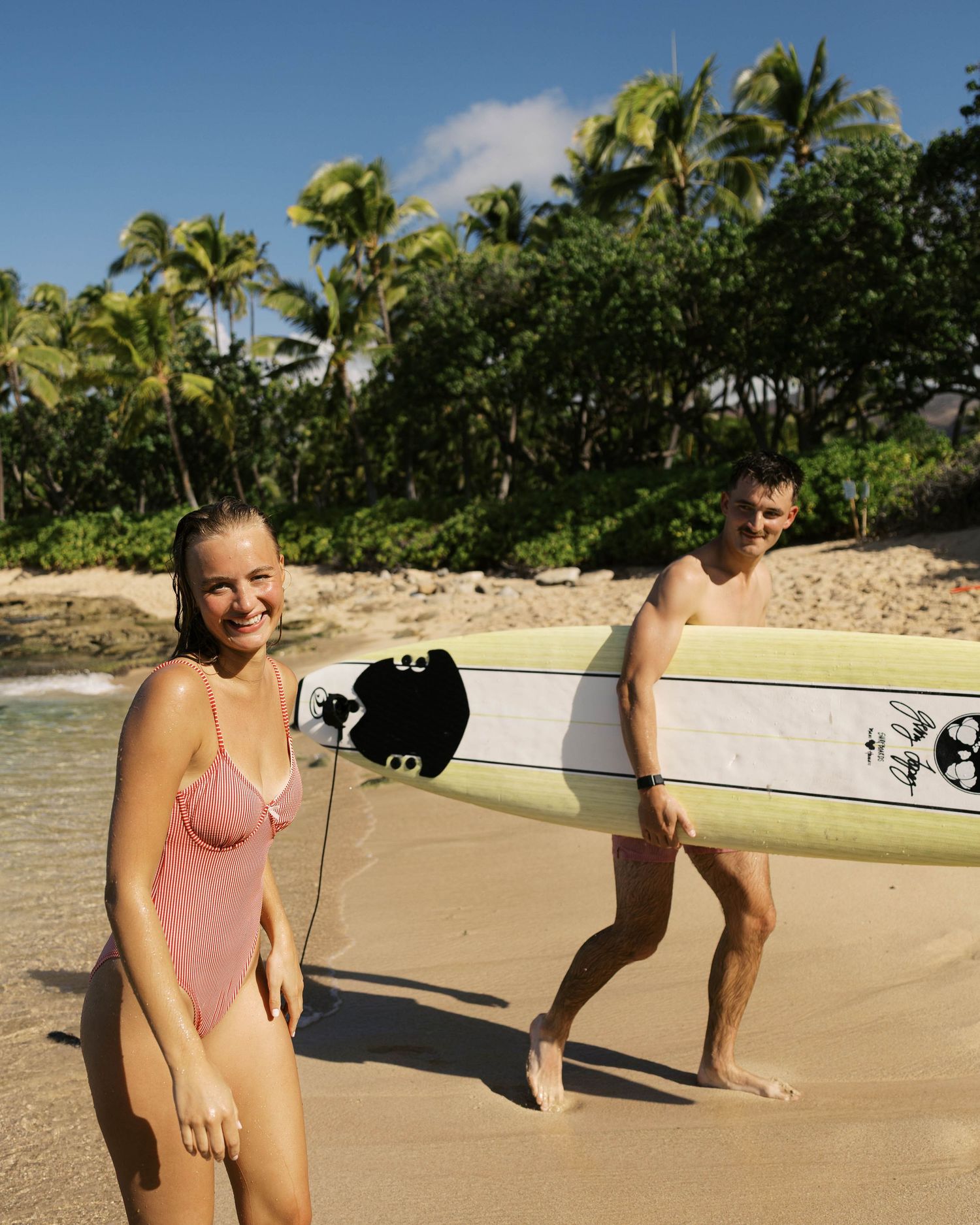 People with surfboards on a sandy tropical beach lined with palm trees.