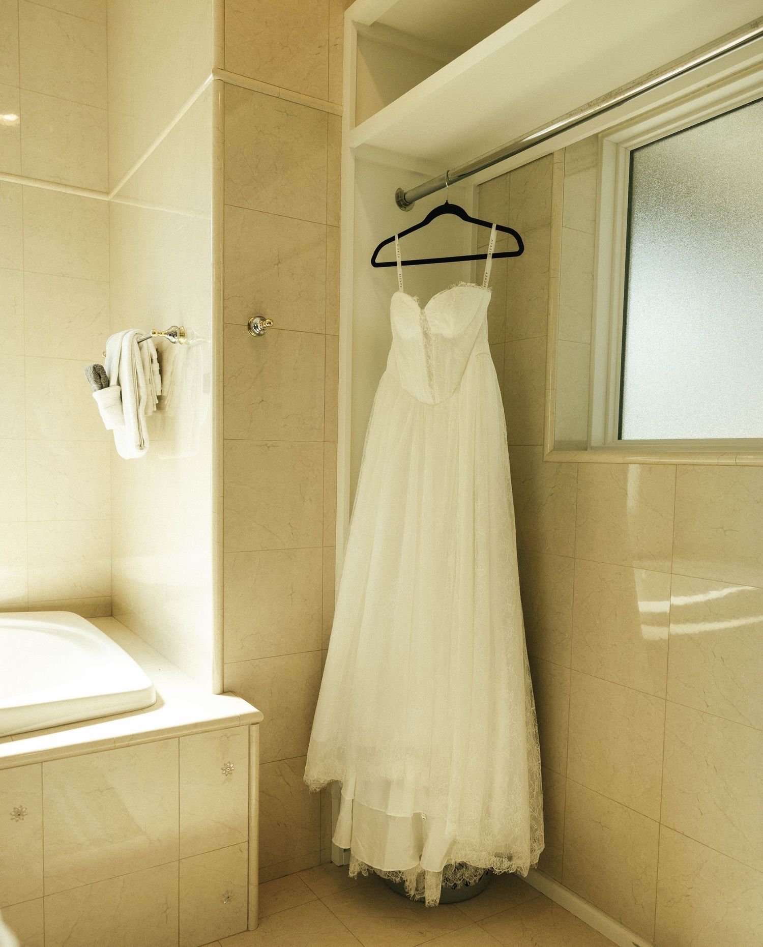 A white wedding dress hangs on a hanger in a beige tiled bathroom.
