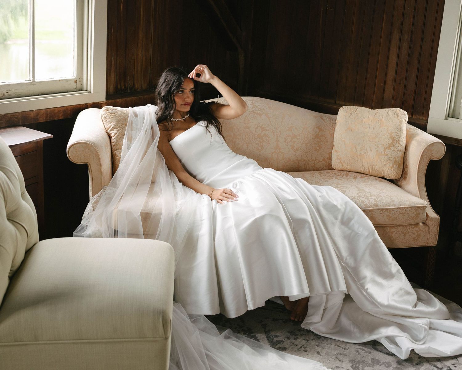 Bride in a white satin gown and veil reclining gracefully on a cream sofa in a rustic wood-paneled room.