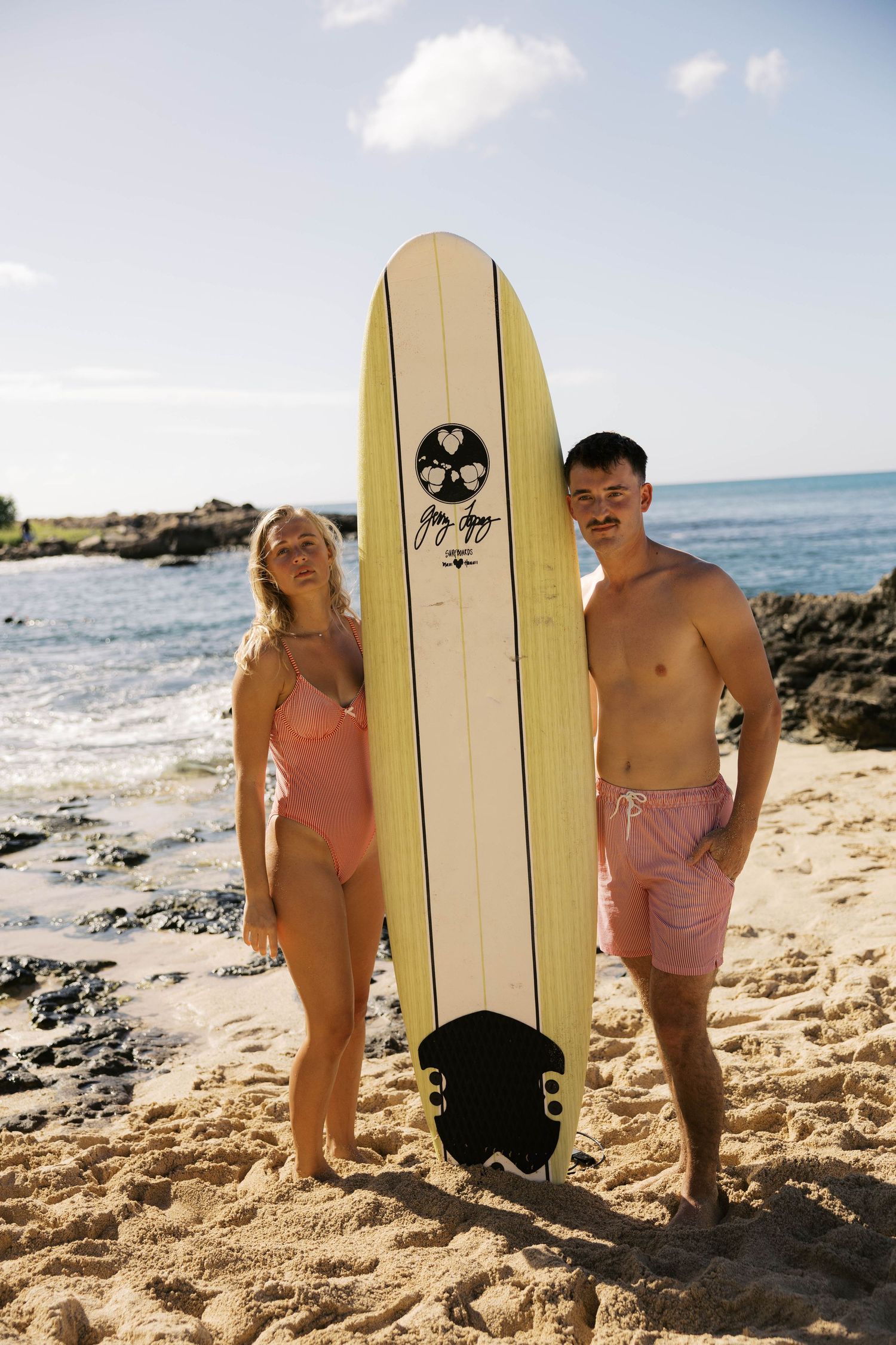 A couple poses with a surfboard featuring a skull logo on a sunny beach with rocky coastline in the background.