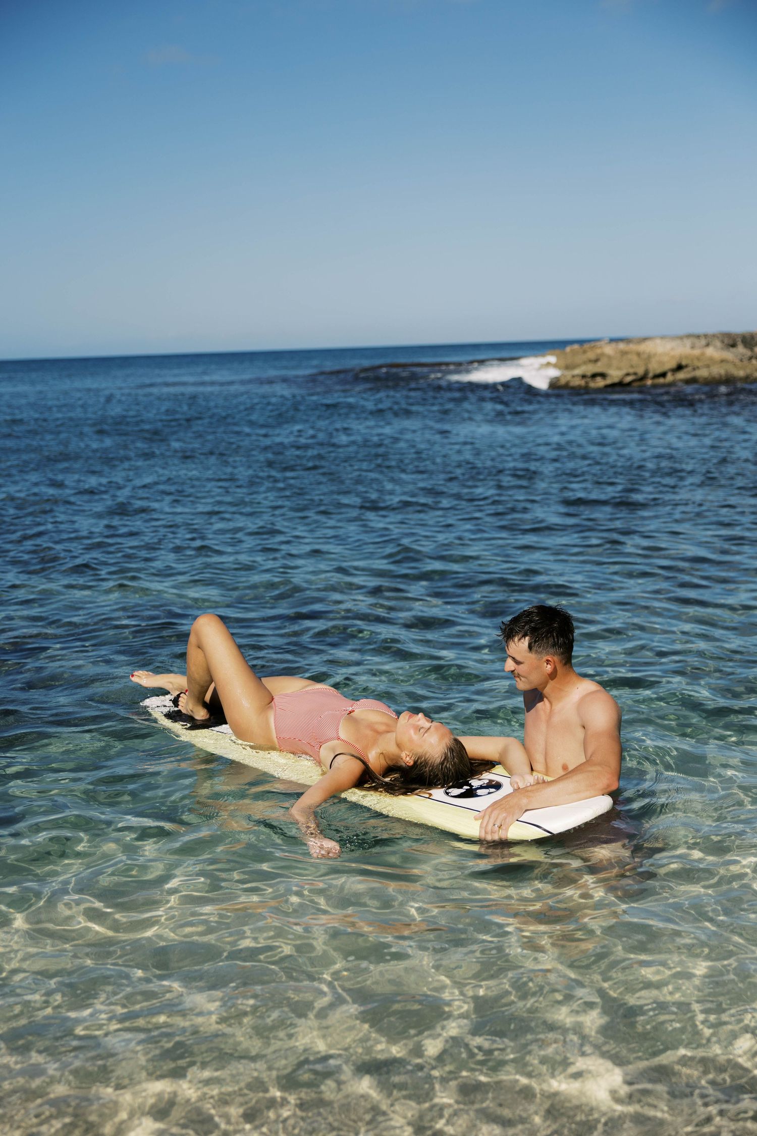 Two people floating on surfboards in shallow tropical waters with rocky coastline and palm trees.
