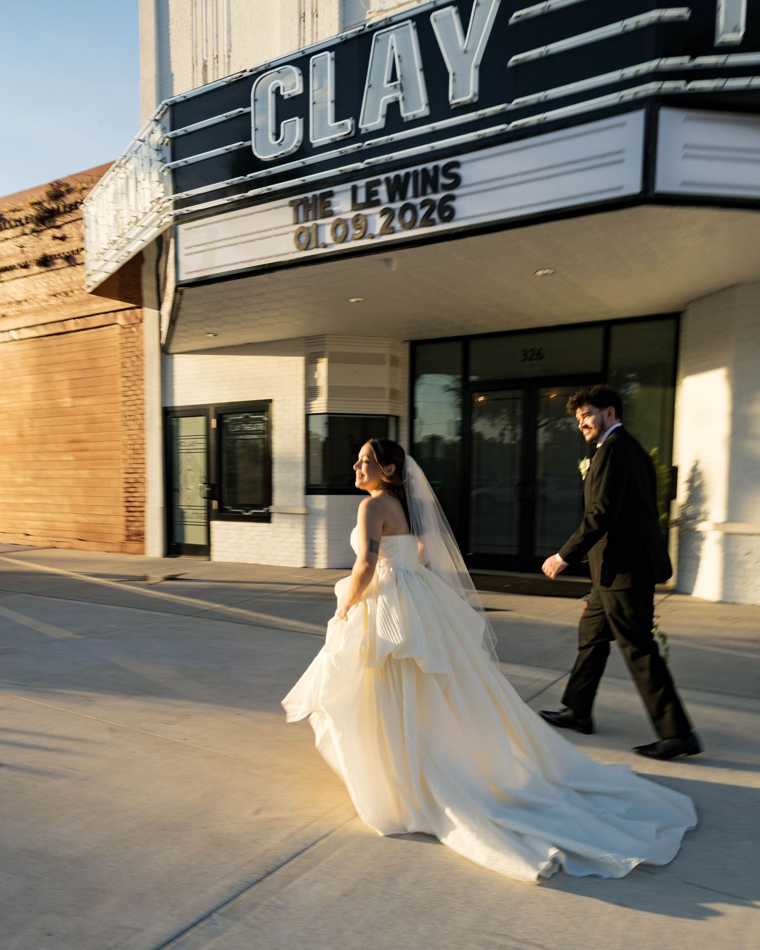 A bride in flowing white gown walks with her groom outside the Clay Theatre.