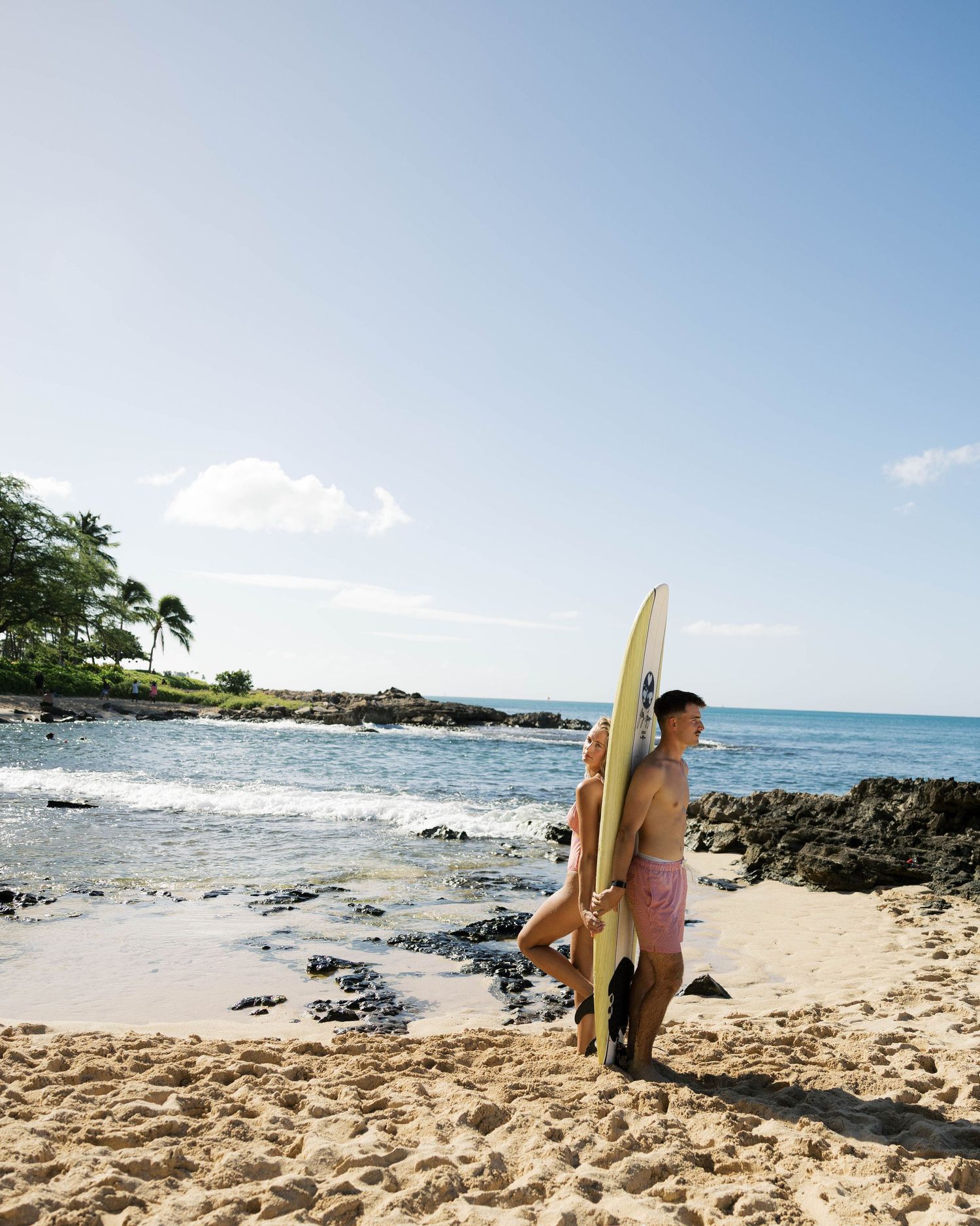 Surfers walking along a scenic beach with volcanic rocks and blue ocean in Hawaii.