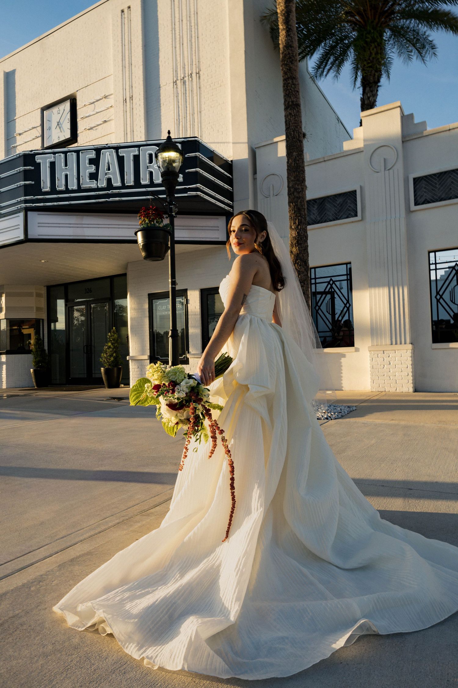 Bride in a classic white gown holds a bouquet while posing outside an Art Deco theater.