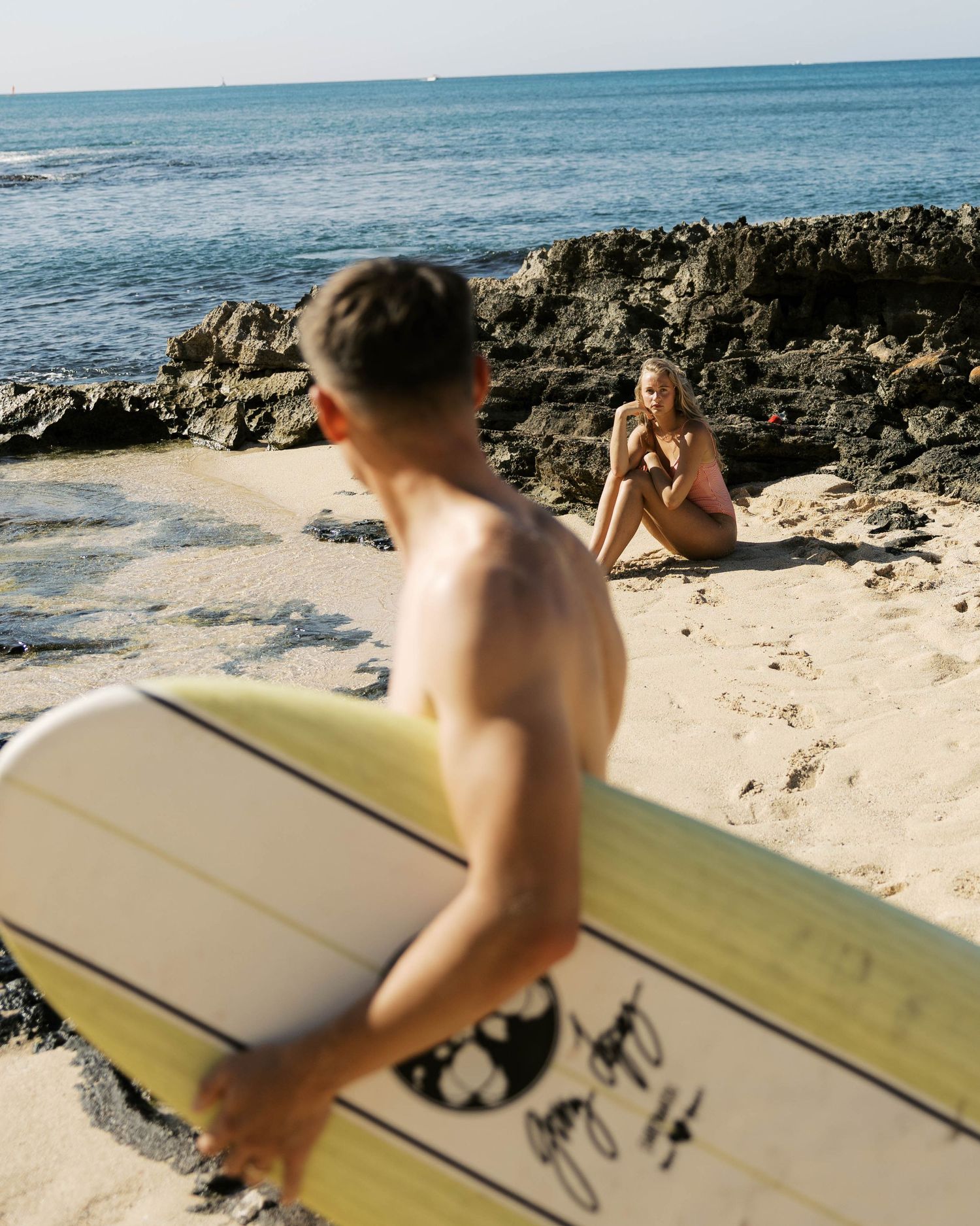 Couple enjoying a sunny day at a tropical beach with surfboard and volcanic rocks.