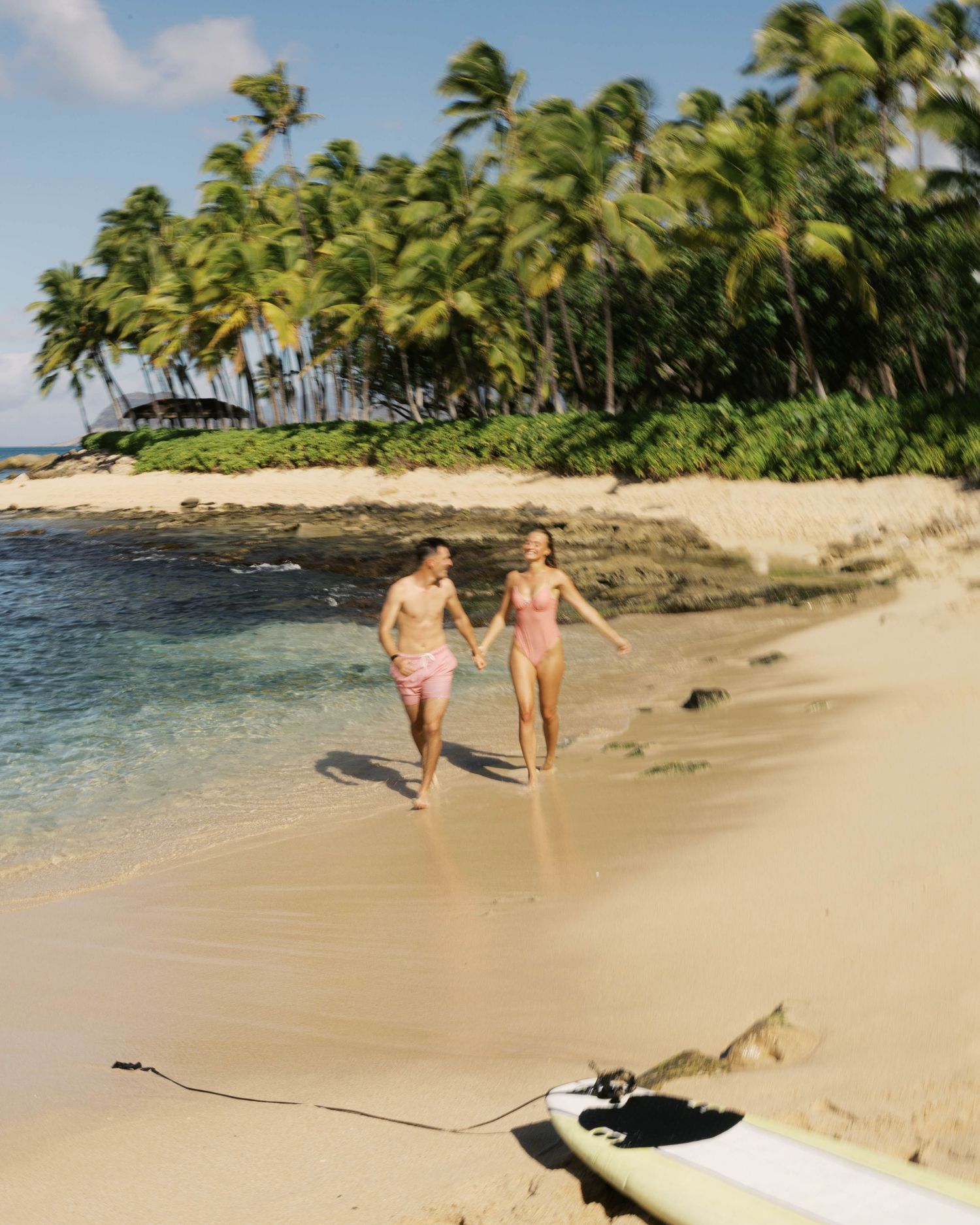 Tropical beach scene with surfers walking along the sand near palm trees and ocean.