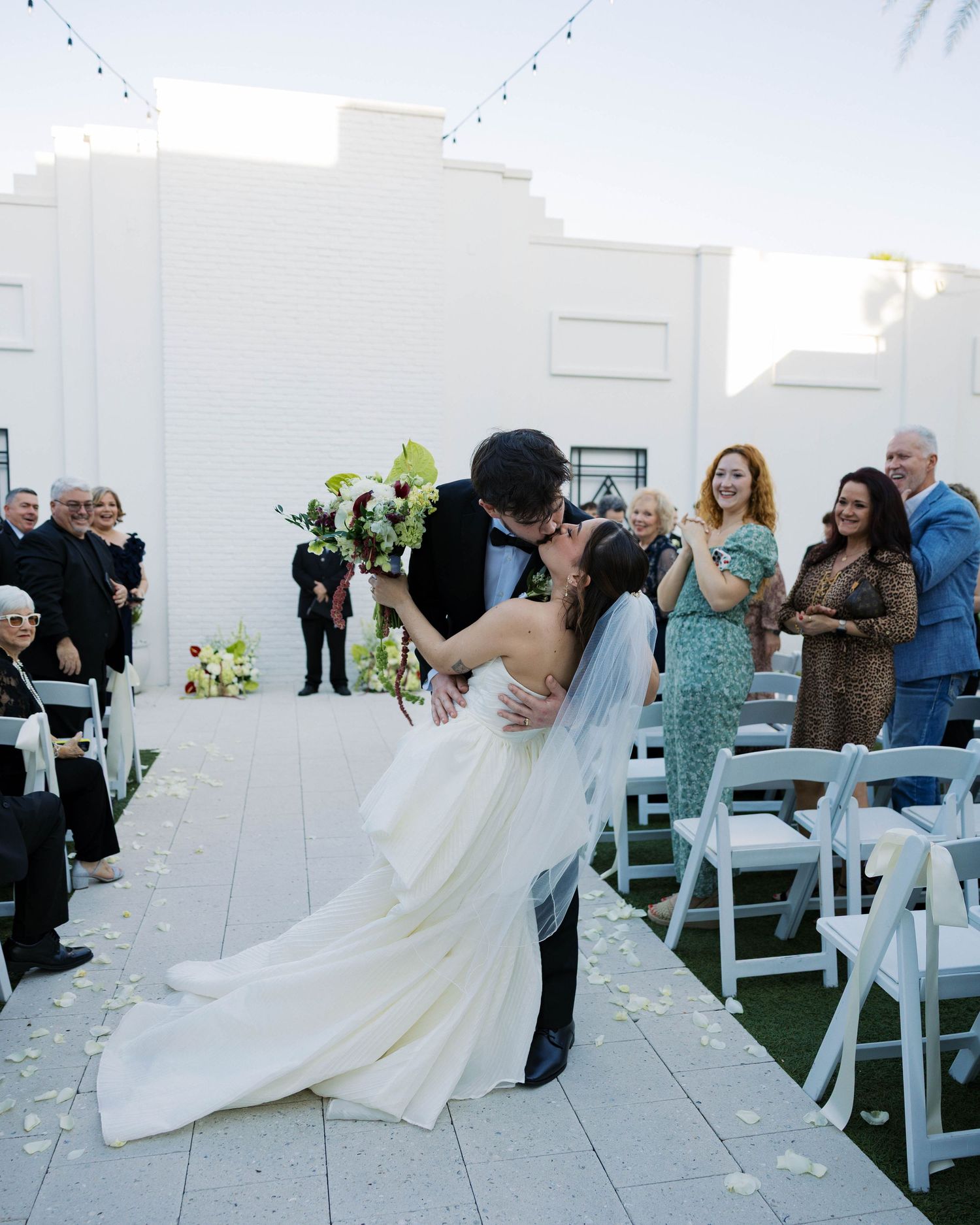 Wedding ceremony candid moments captured against white architectural backdrop with string lights.