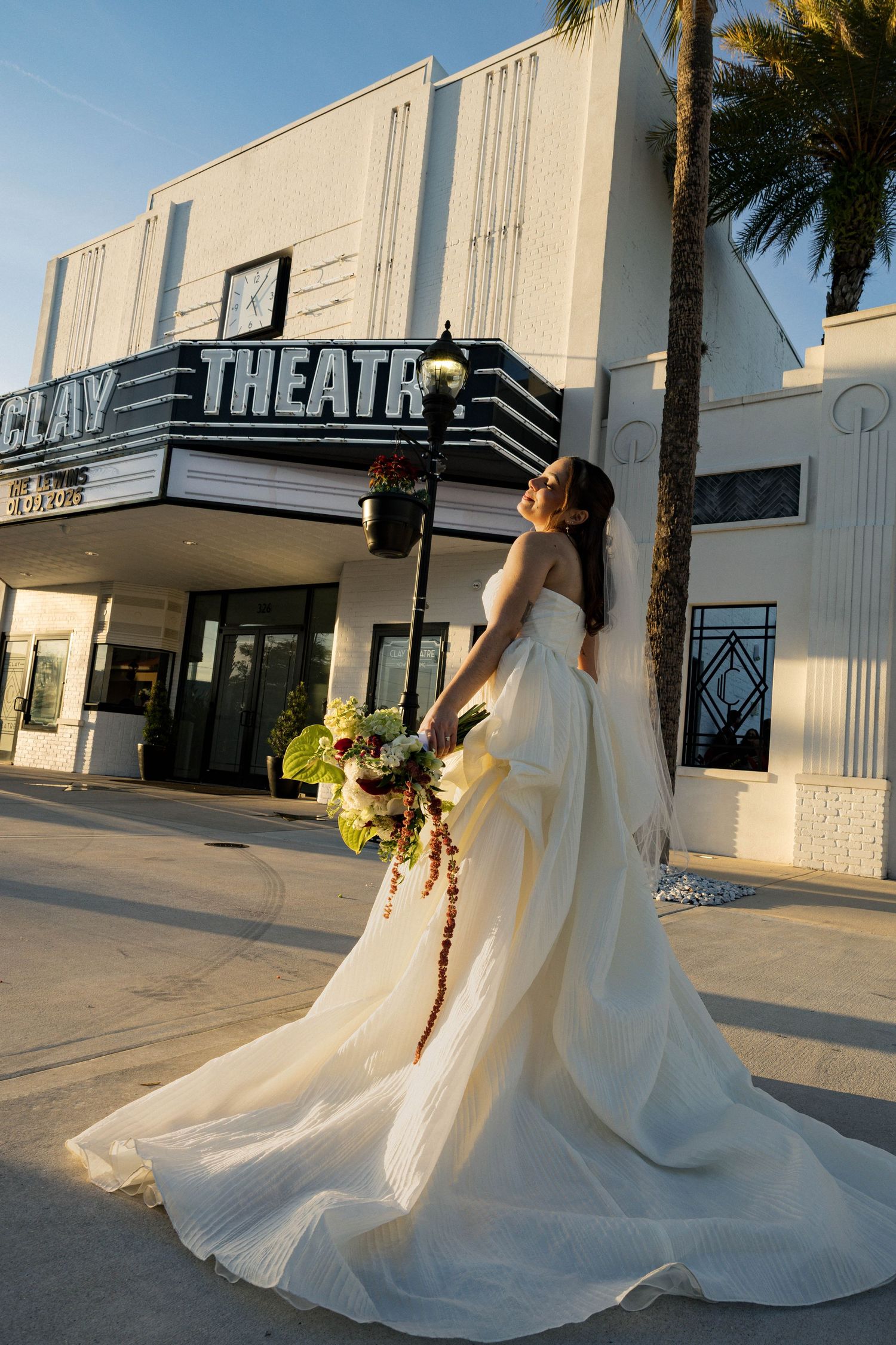 Bride in a dramatic white ballgown poses with her bouquet outside a historic theater.