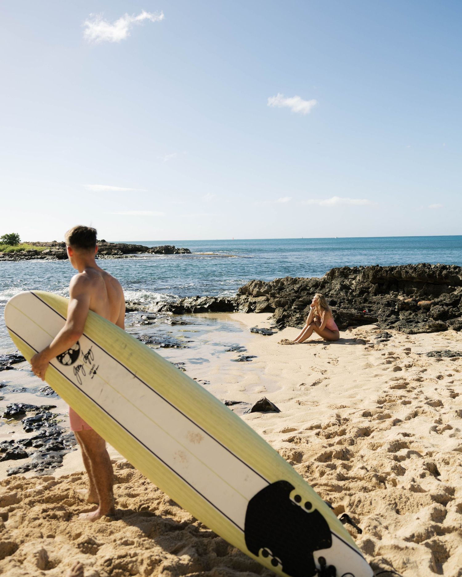 Beach scene with surfer holding board while standing on sandy shore near volcanic rocks.
