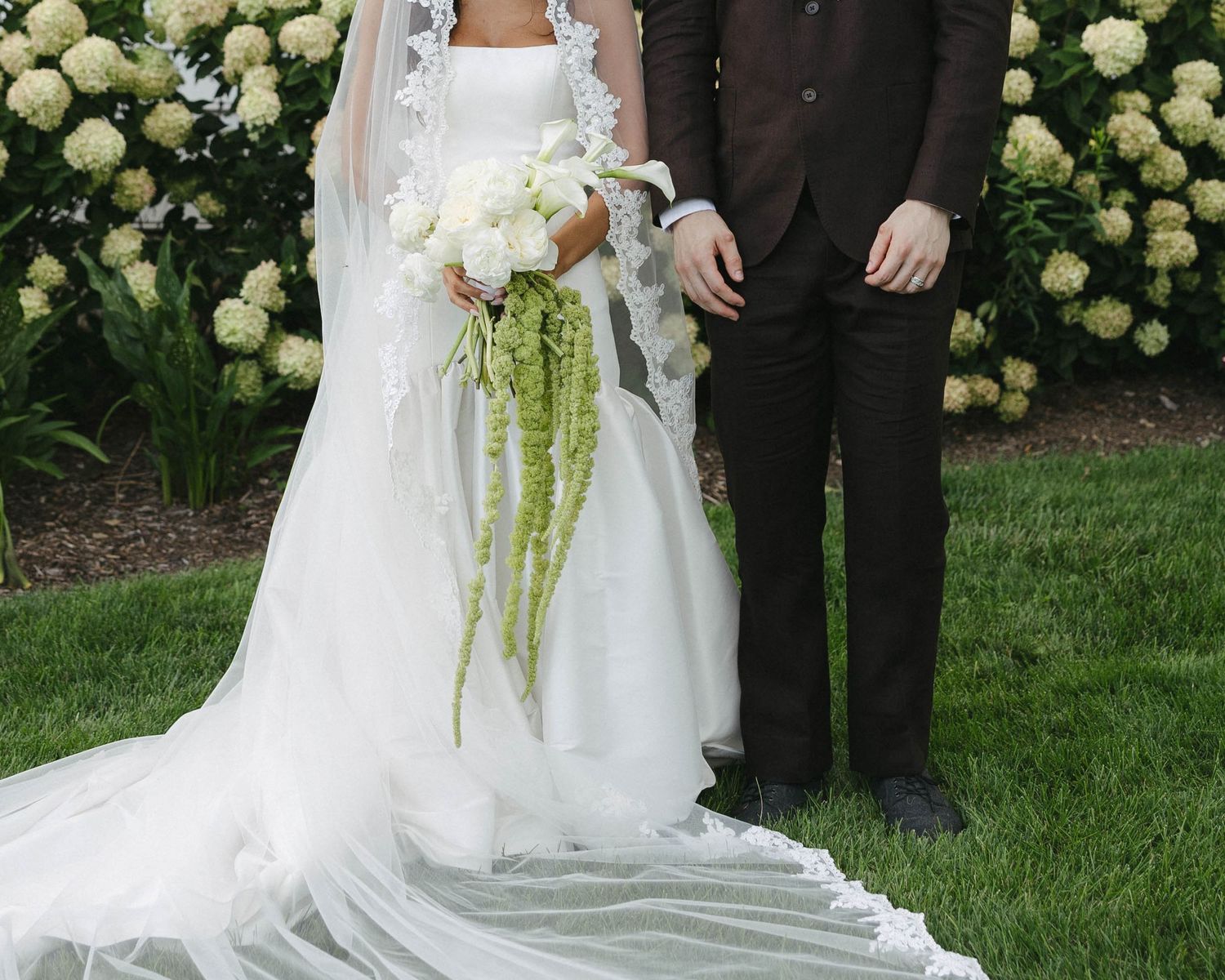 Bride and groom standing outdoors on green grass, bride wearing white gown with veil and holding white and green bouquet.