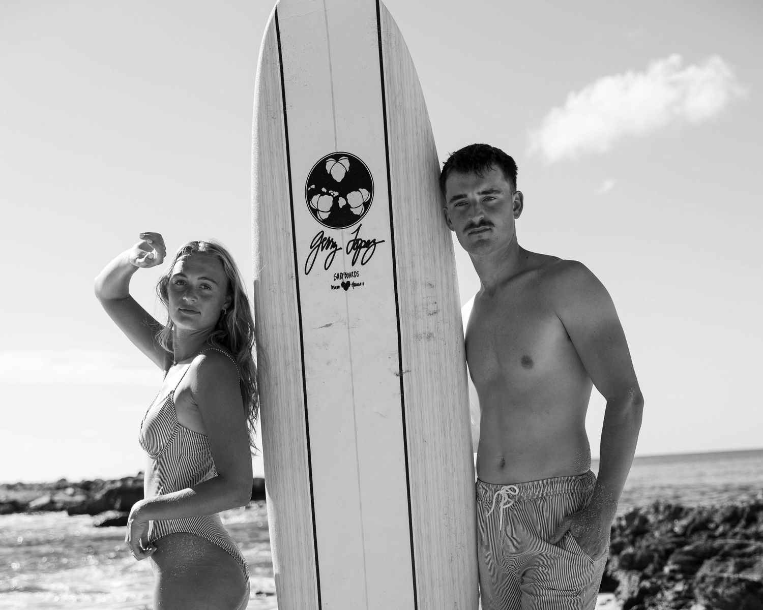Black and white beach portrait with vintage surfboard against coastal background.