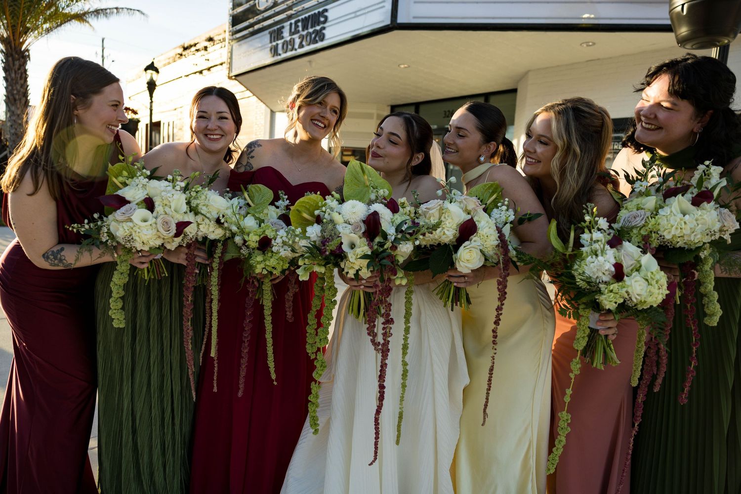 A wedding party with bridesmaids in burgundy and green dresses holding elegant white floral bouquets.