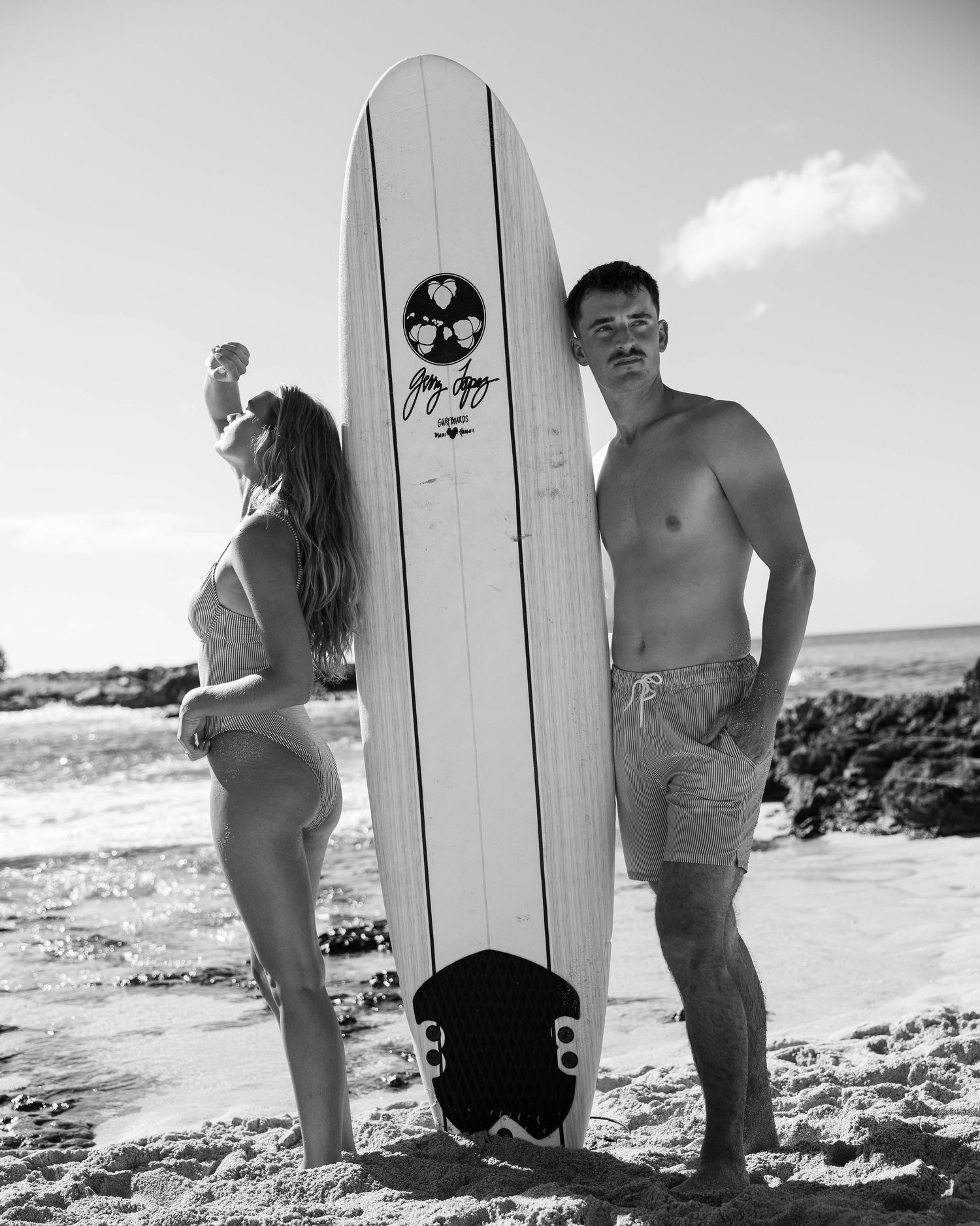 Black and white beach portrait with surfboard and rocky coastline.