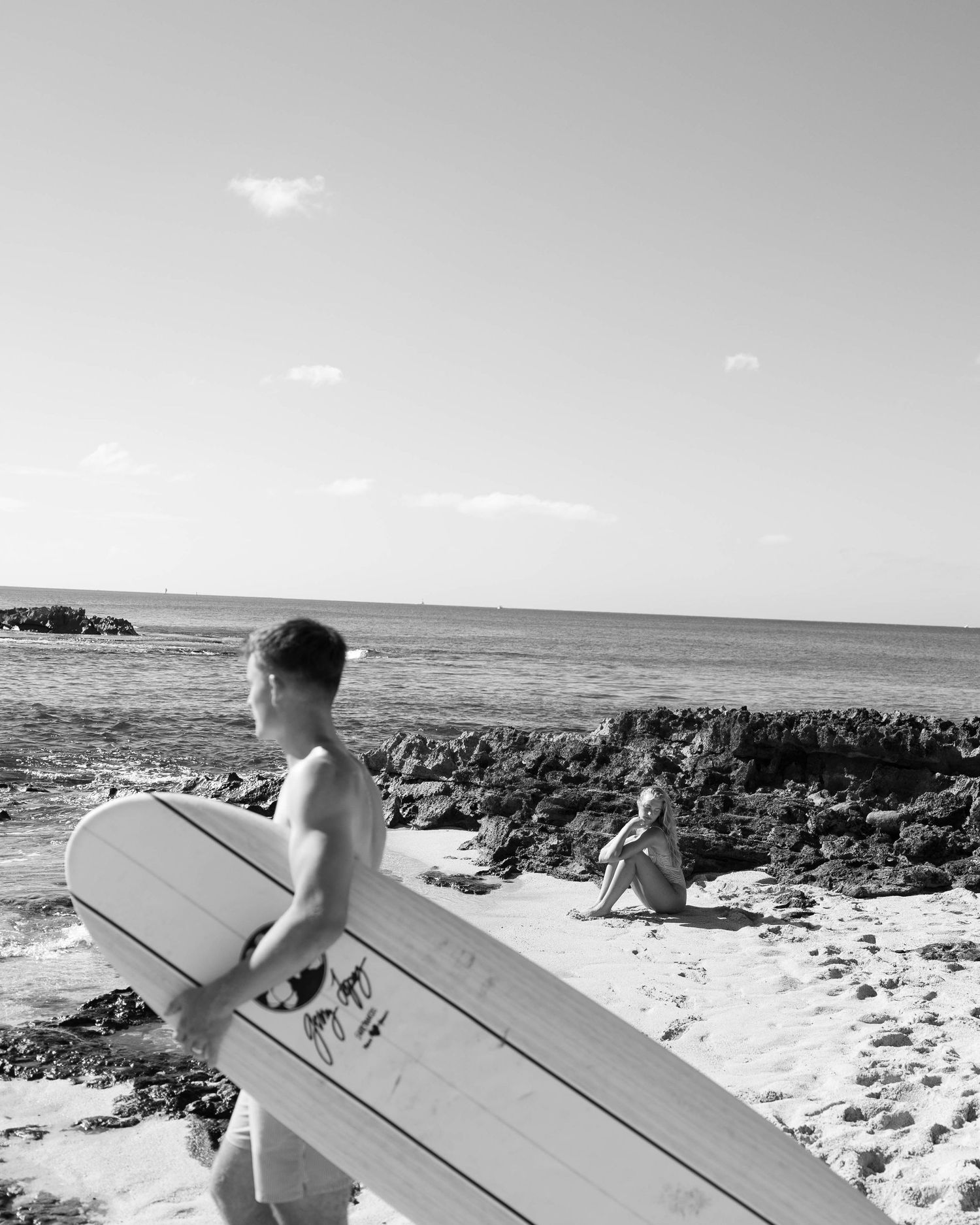 Black and white photo of surfers carrying board across rocky shoreline.