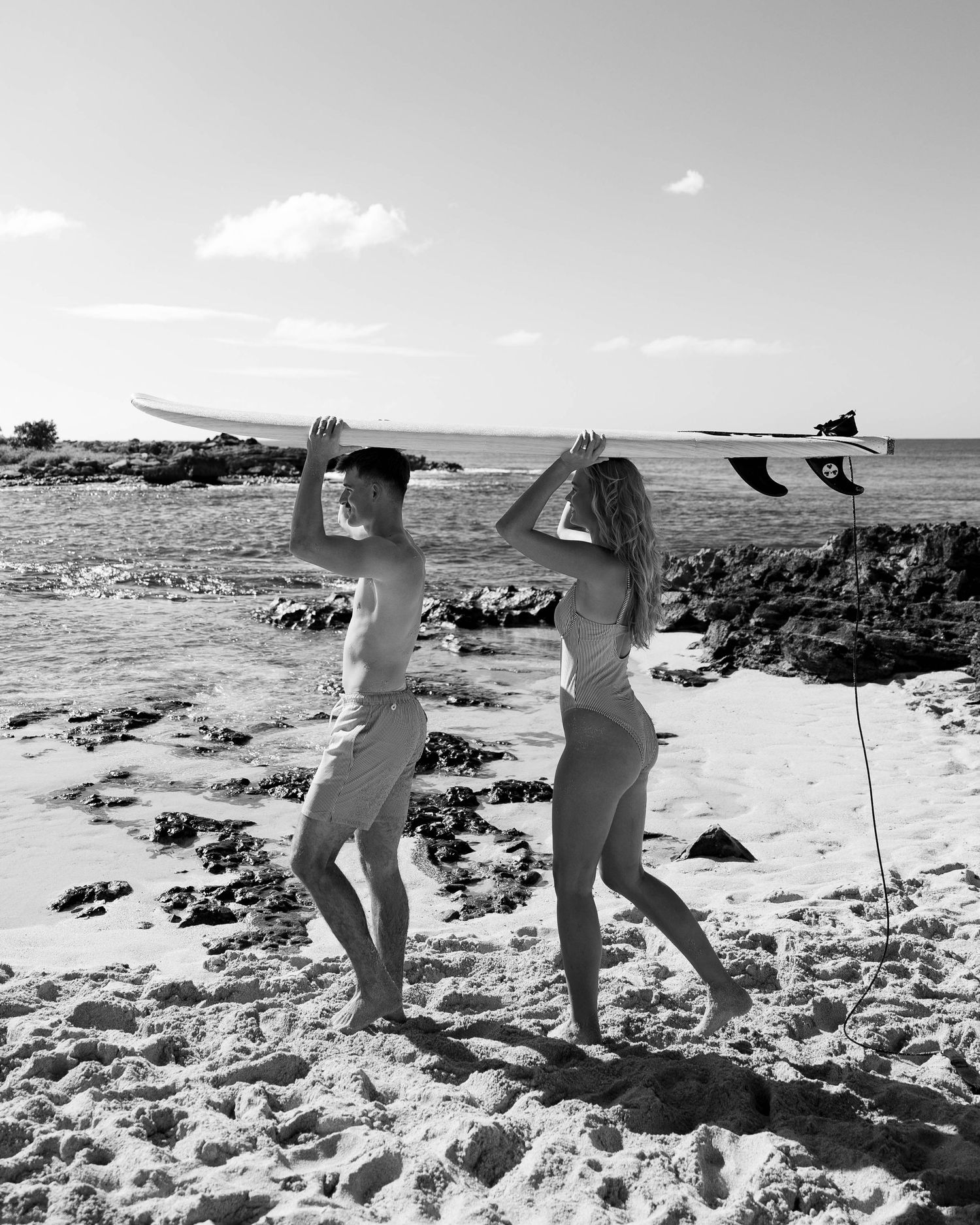 Black and white sequence of people carrying surfboard across sandy beach.