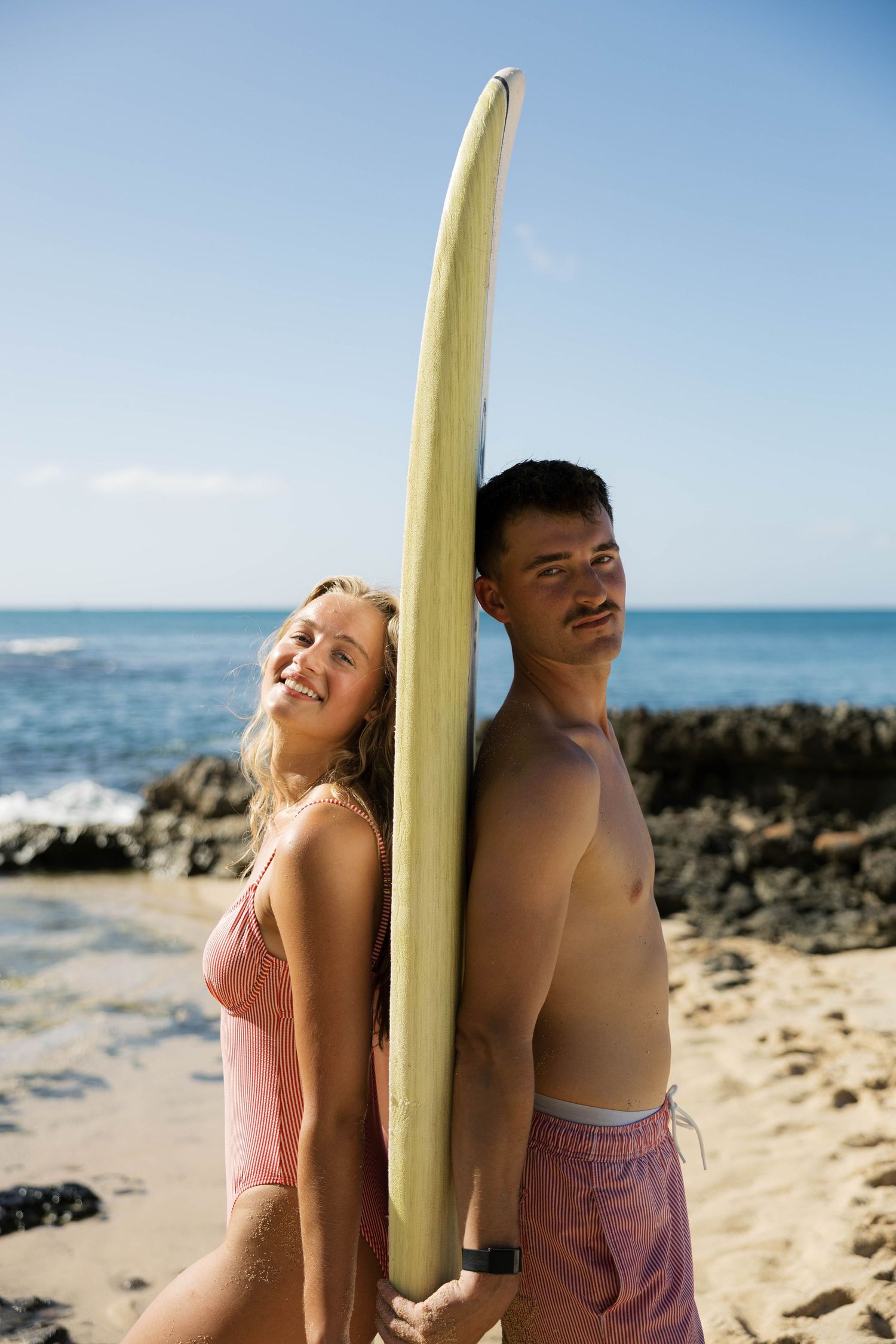 Couple in beachwear posing with surfboard against rocky coastline.