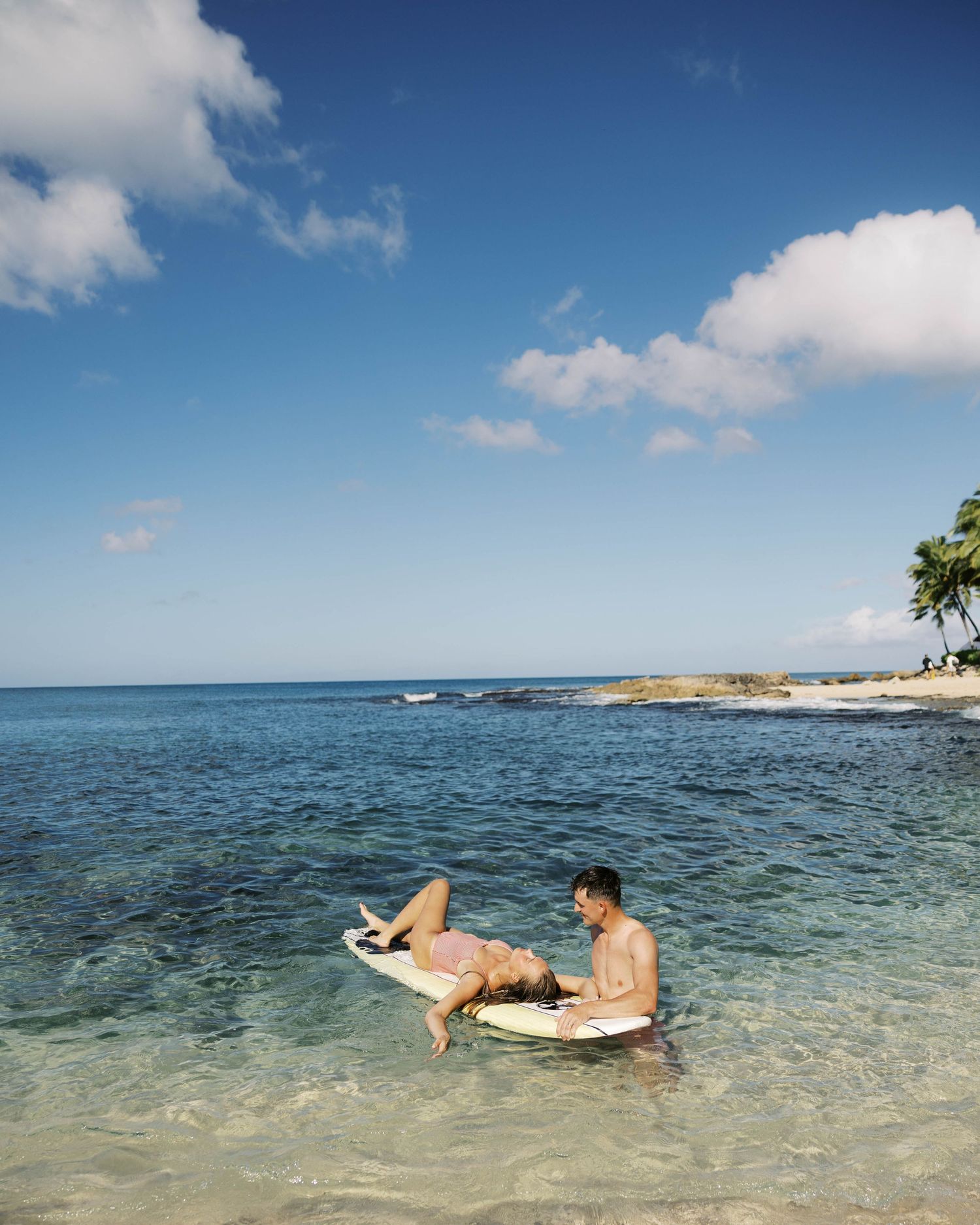 Surfers floating on boards in tranquil blue ocean water near a scenic beach coastline.
