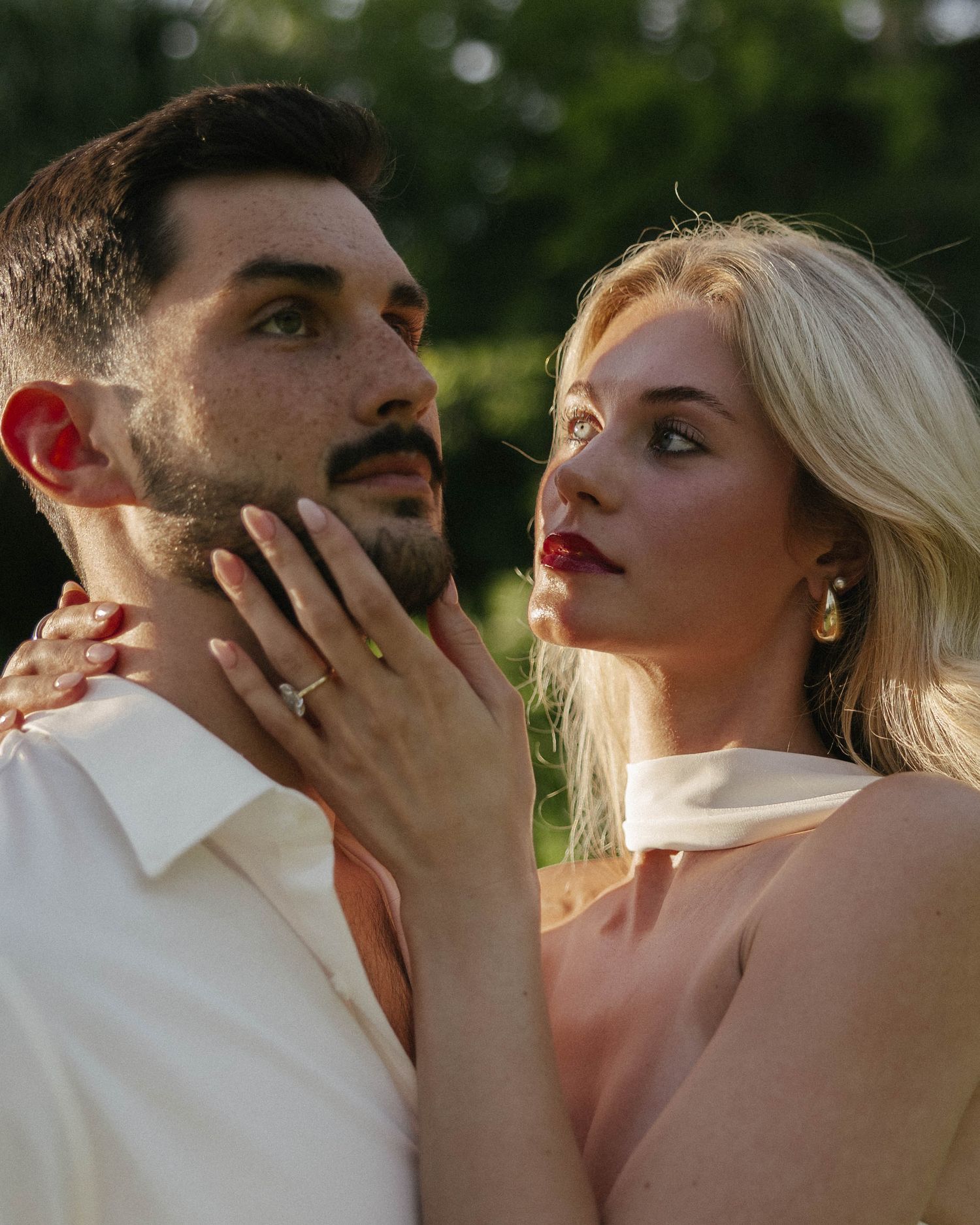 Romantic couple portrait with blonde woman in elegant earrings touching bearded mans chin outdoors at golden hour.