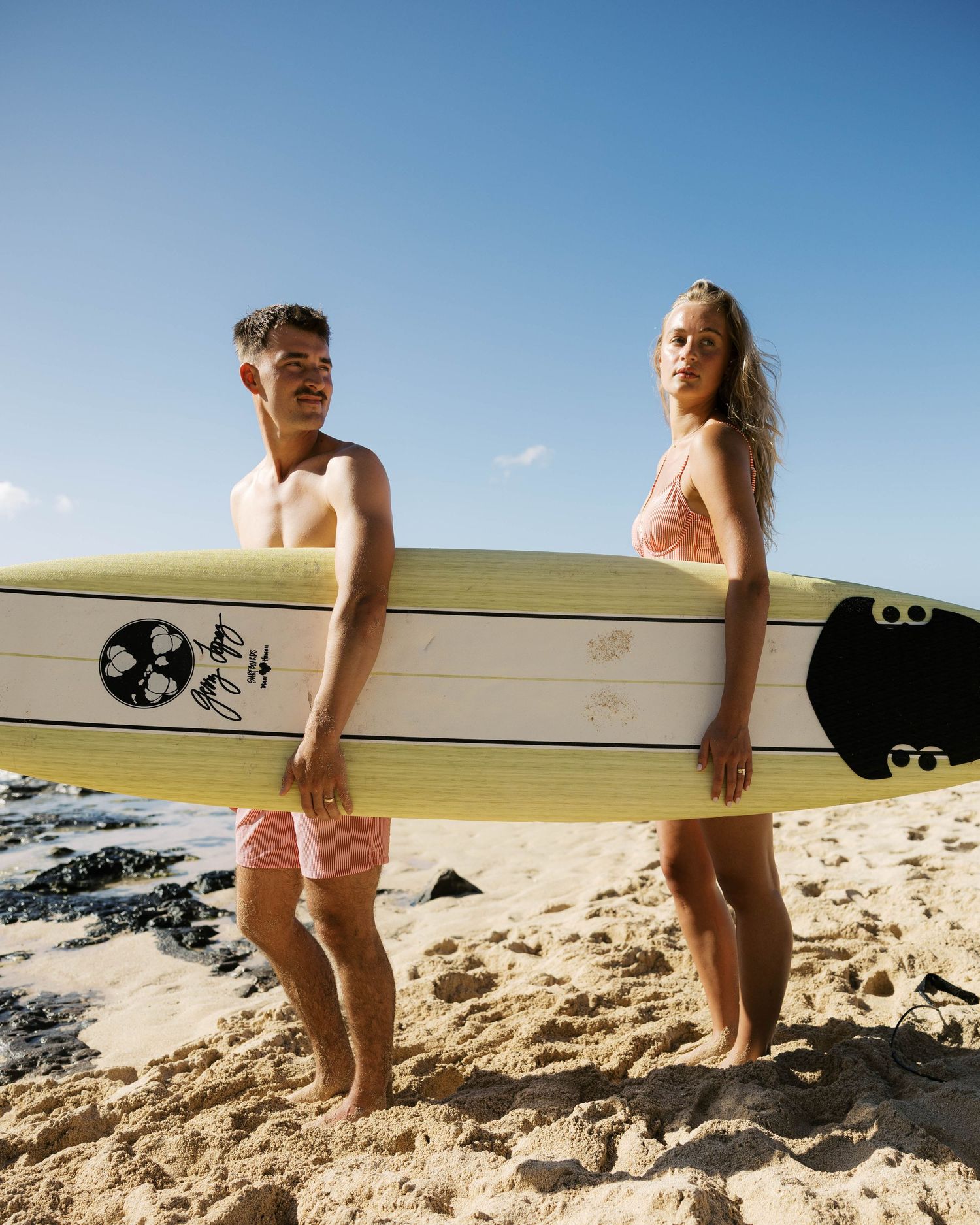 Beachgoers carrying surfboard across sandy shore on sunny day.