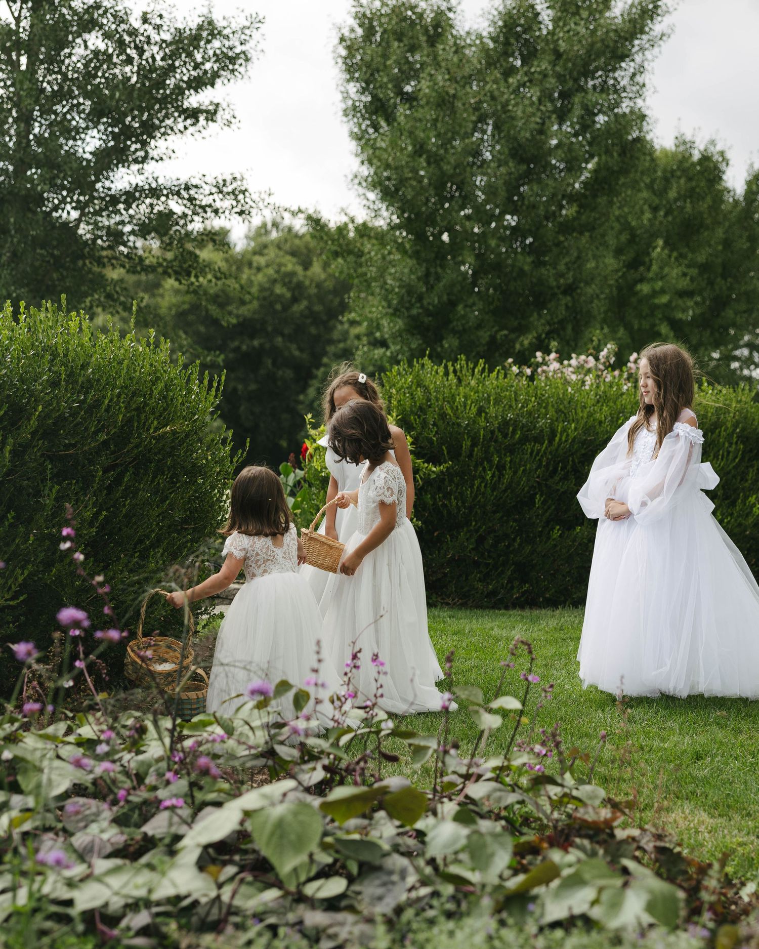 Young flower girls in white dresses gather in a lush garden, one holding a wicker basket, during a wedding celebration.